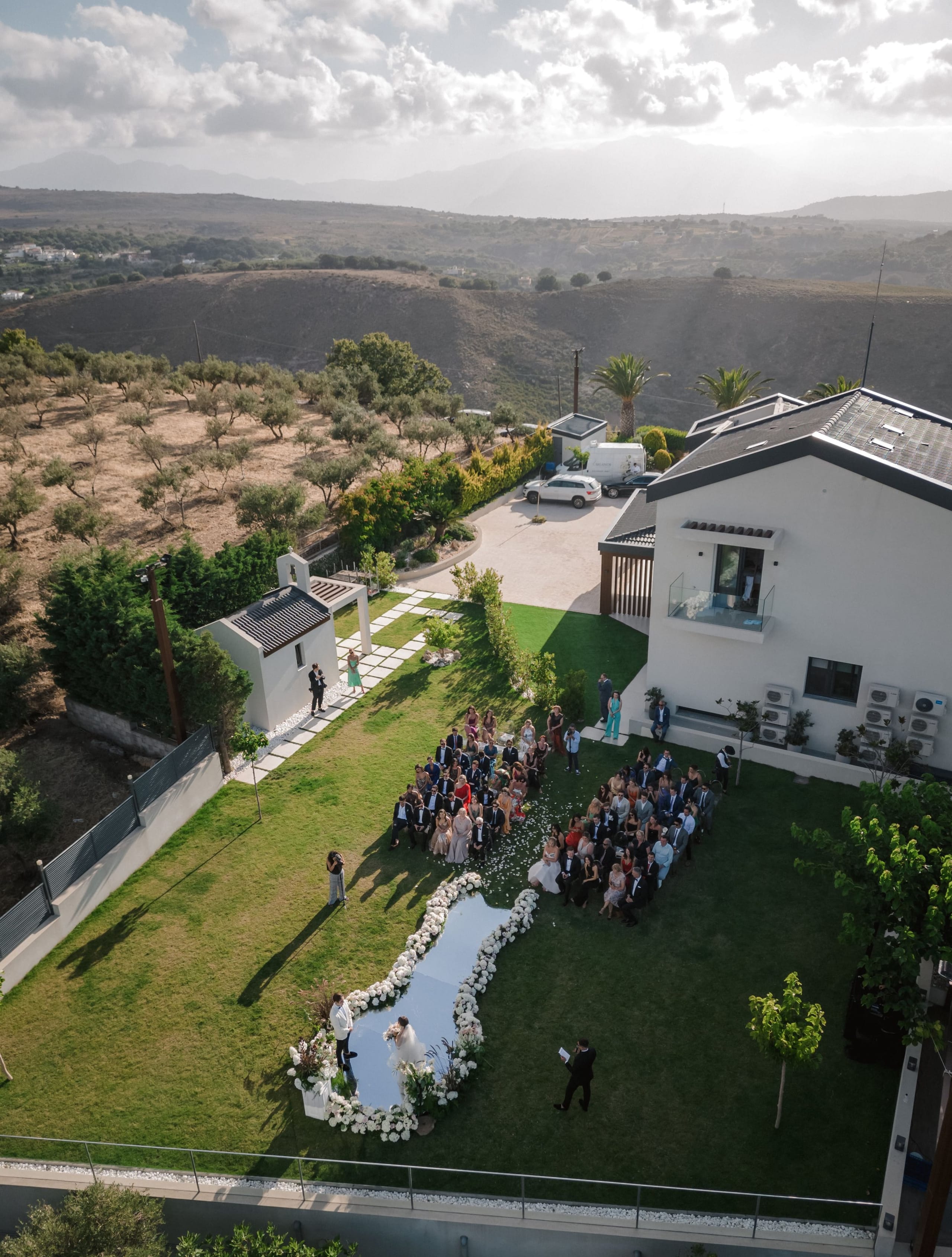 Aerial view of a wedding ceremony on a lawn at Amphi Residence in Tria Monastiria, Crete, Greece, with guests seated in rows facing a small pool bordered by a curving white floral arrangement of roses and greenery. A bride in a white gown and a groom in a suit stand at the water's edge, while a celebrant in dark clothing faces them. The white villa, olive groves and rolling hills under a soft, hazy sky complete the scene.
