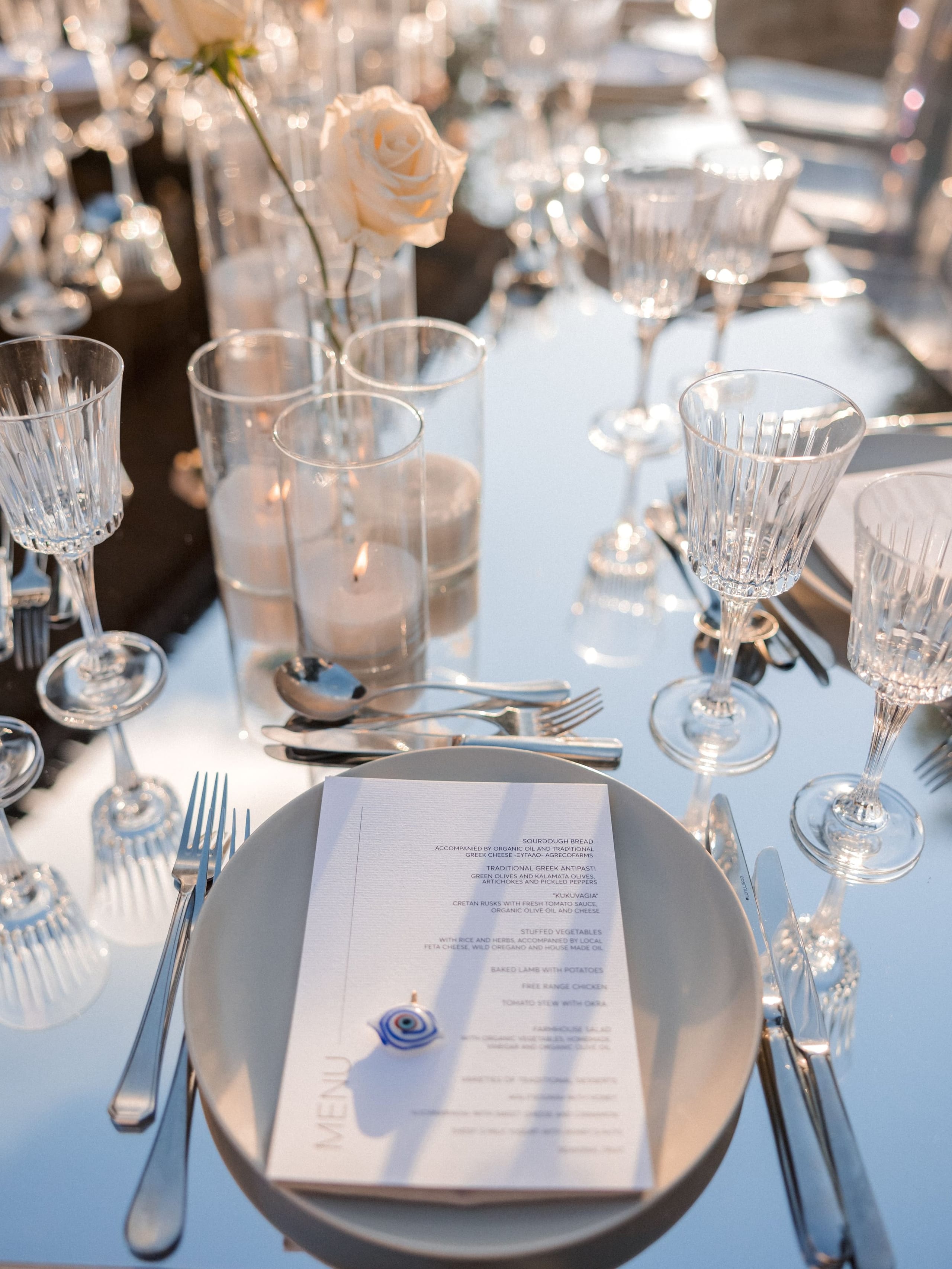 A wedding table setting at Amphi Residence in Tria Monastiria, Crete, Greece, dressed in cream garden roses rising from clear glass cylinders with softly lit candles. A white menu card with a small blue evil-eye charm rests on a pale grey plate, flanked by polished silver cutlery and tall ribbed crystal glasses. The mirrored, pale blue-grey table reflects the warm late-afternoon light, giving the scene a calm, elegant and airy mood.