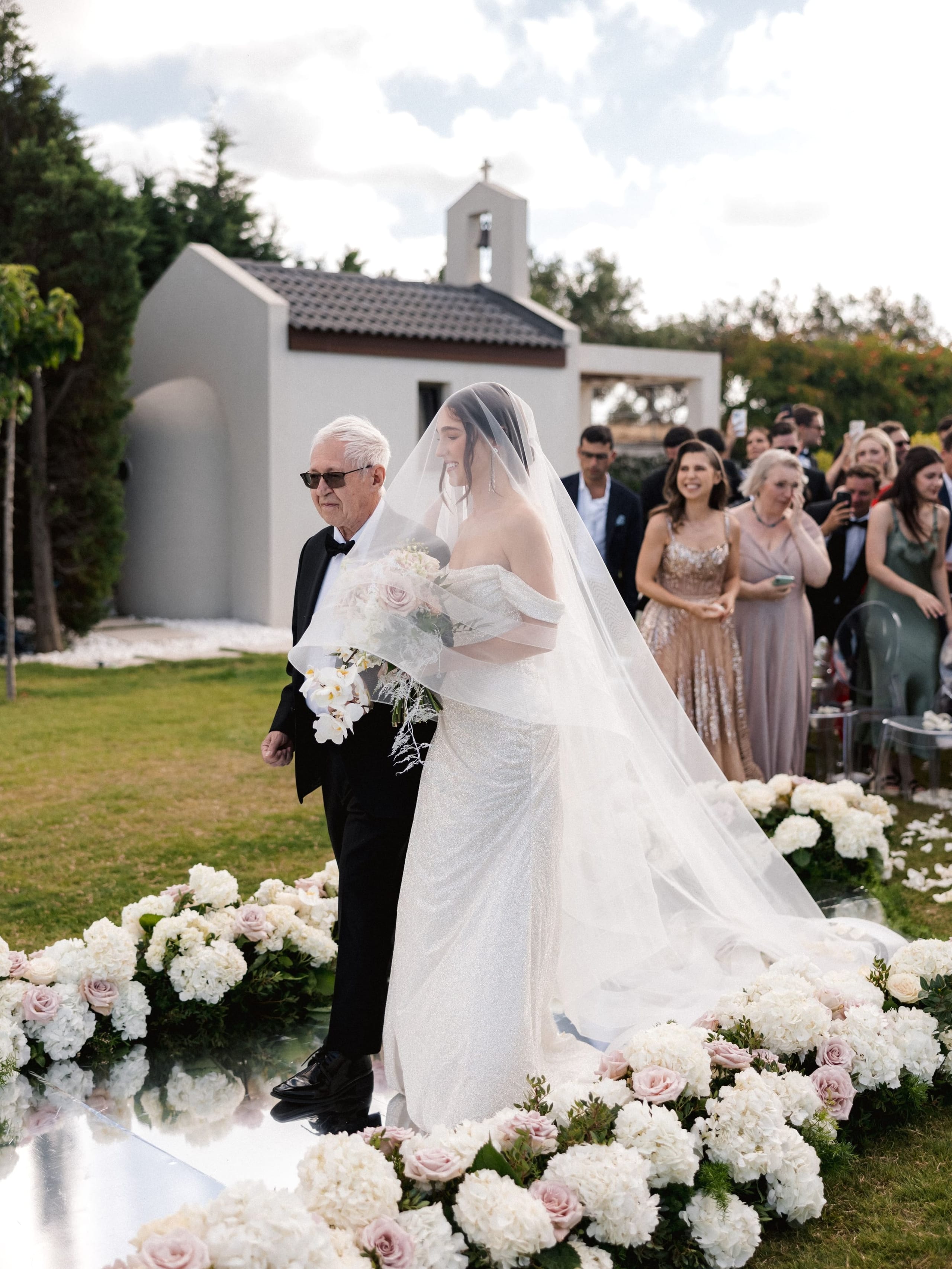 A bride in a sparkling off-the-shoulder gown and long sheer veil walks down an aisle lined with white hydrangeas, pale pink garden roses and white orchids, set on a mirrored runner, while an older man in a black tuxedo and sunglasses walks beside her holding her arm. Behind them guests in dusty pink and sage dresses smile and raise their phones, with a small white chapel under a bright sky. This is a wedding ceremony aisle at Amphi Residence in Tria Monastiria, Crete, Greece.