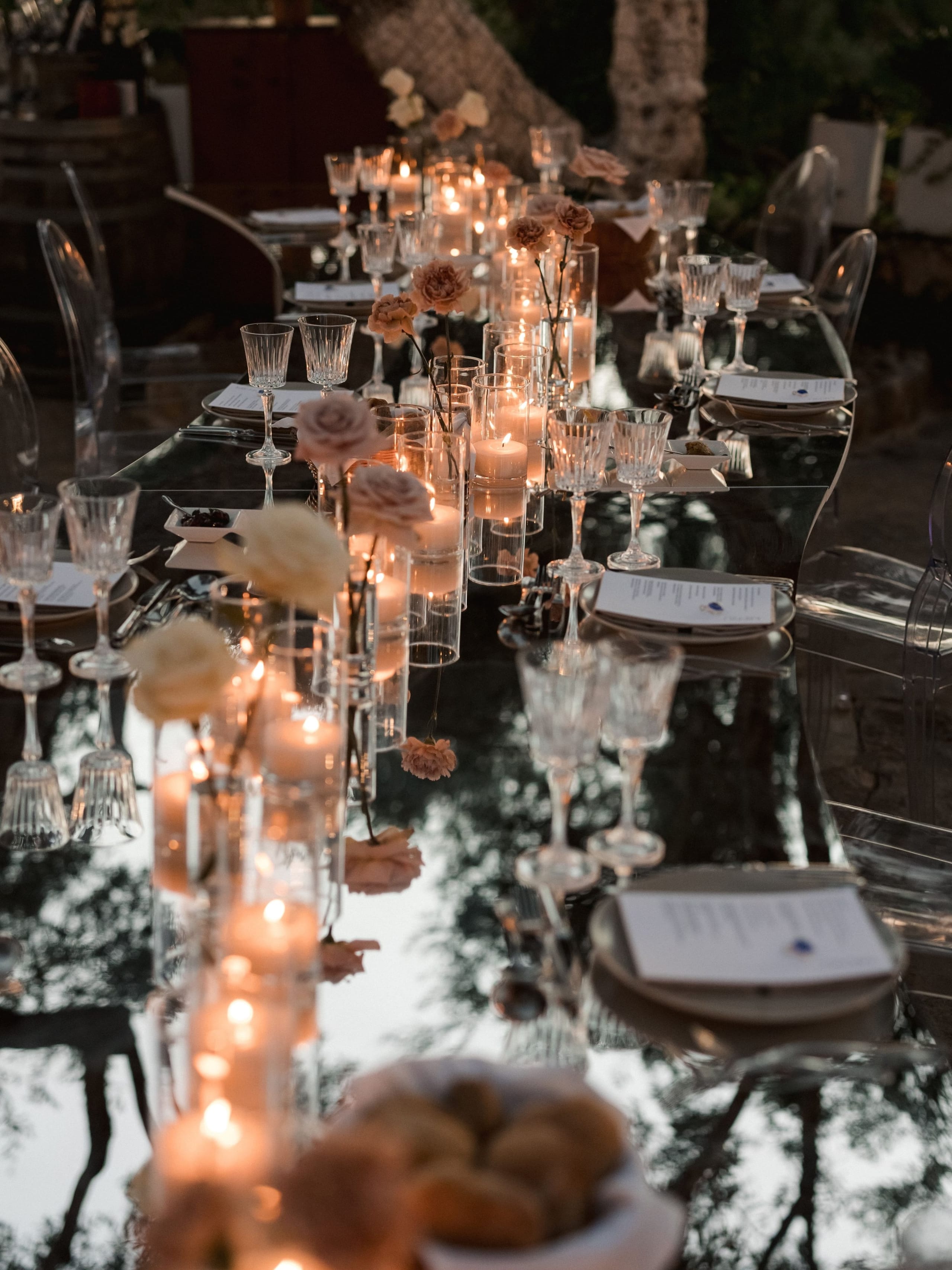 A long glass-topped wedding dining table at dusk, lined with clusters of lit pillar candles in clear glass cylinders and dusty-peach garden roses and cream ranunculus in tall stems. Crystal wine glasses and printed menu cards sit at each place, reflecting the candlelight and the dark trees above. This is a wedding table setting at Amphi Residence in Tria Monastiria, Crete, Greece, with a warm, intimate mood.