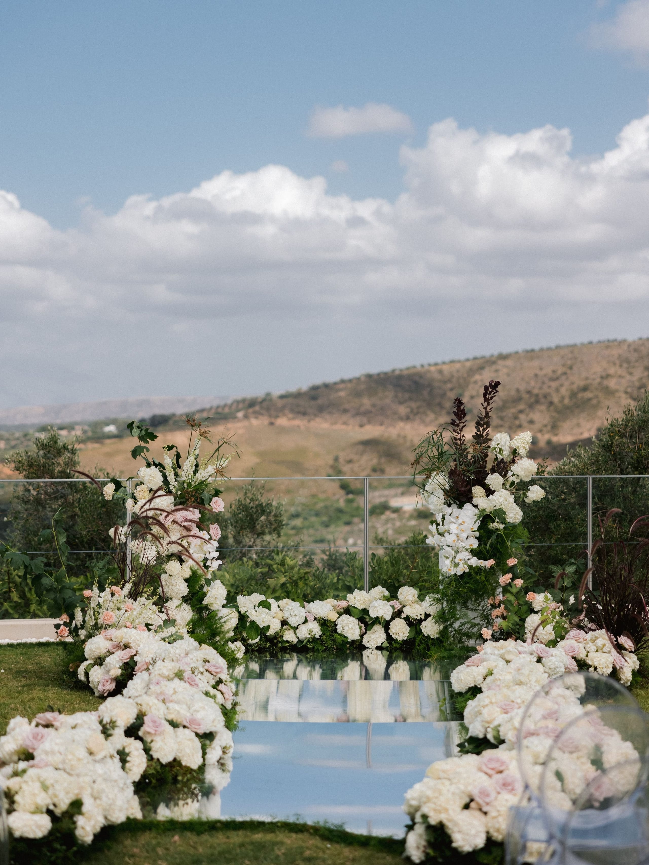 A wedding ceremony aisle in Greece, lined on both sides with dense white hydrangeas and blush roses, leading down a mirrored walkway toward an arch of white orchids and tall dark grasses at Amphi Residence in Tria Monastiria, Crete. Beyond the terrace glass rail, olive trees and golden, sun-dried hills roll away under a pale blue sky with soft white clouds.