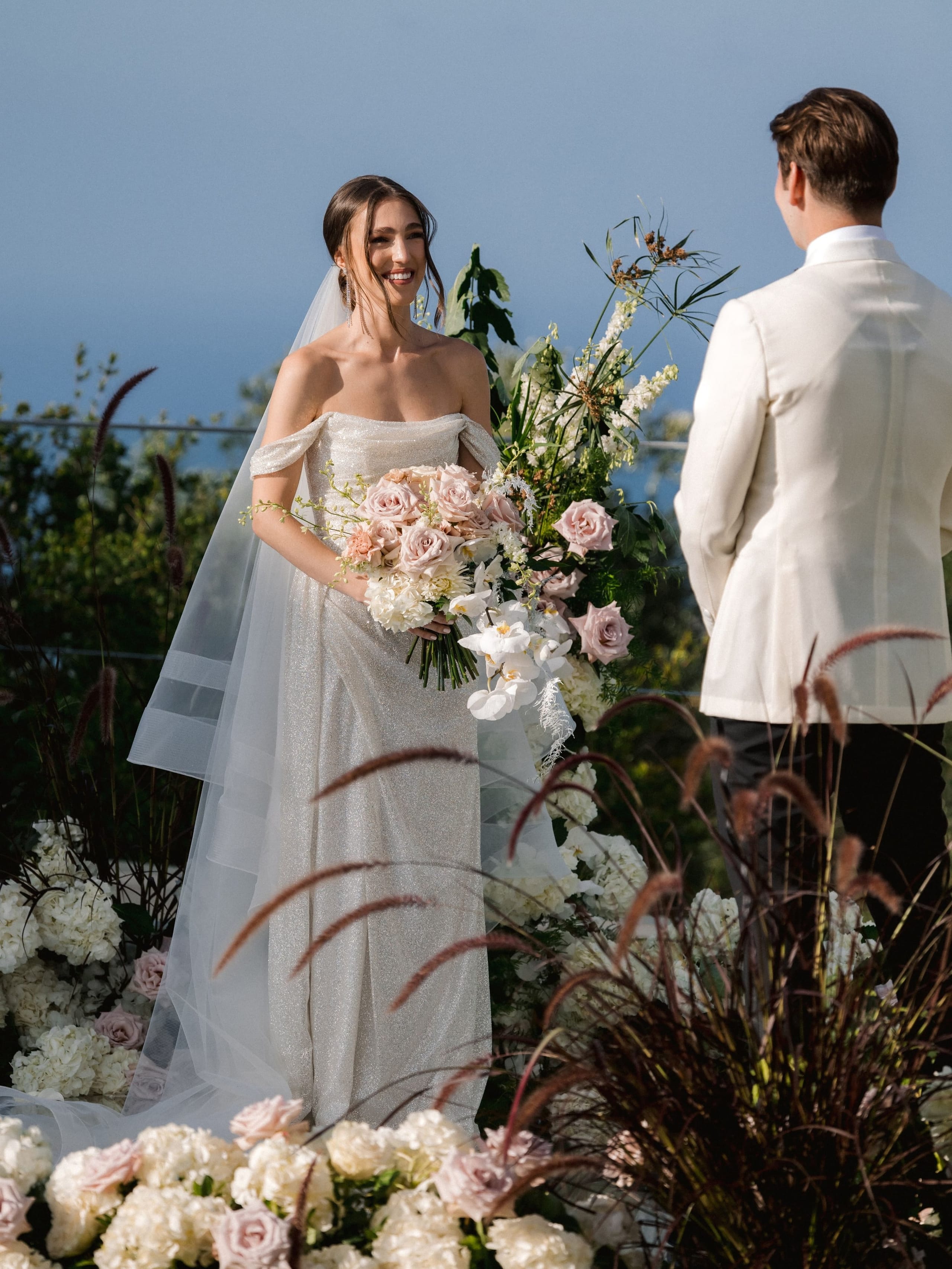 A smiling bride in a shimmering off-the-shoulder gown and sheer veil stands holding a loose bouquet of blush garden roses, white orchids and hydrangeas, while a groom in a cream jacket faces her from the right. Behind them rise tall white delphinium-like spikes and dark plumes of grass, with hydrangeas and pale pink roses banking the foreground, all set against a clear blue sky. These are wedding flowers for a ceremony at Amphi Residence in Tria Monastiria, Crete, Greece.