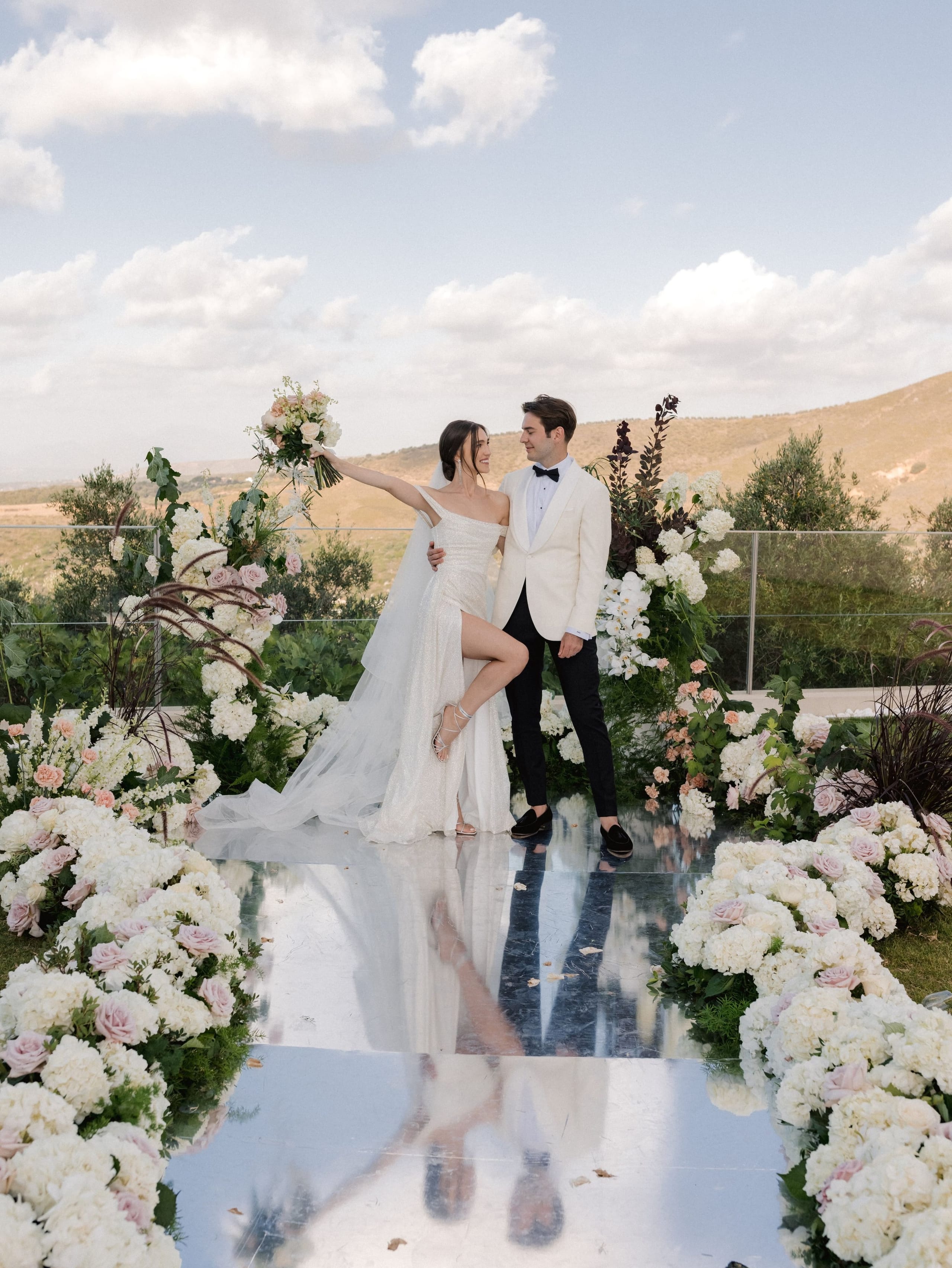 A bride in a sparkling one-shouldered gown and a long veil stands on a mirror-polished aisle, lifting a cream and blush bouquet high while she smiles at the groom in an ivory dinner jacket and black bow tie. Both sides of the aisle are lined with dense white hydrangeas and pale pink roses, with tall arches of white blooms and dark plumes behind them. This wedding flower display at Amphi villa in Greece is set against olive trees and dry golden hills.