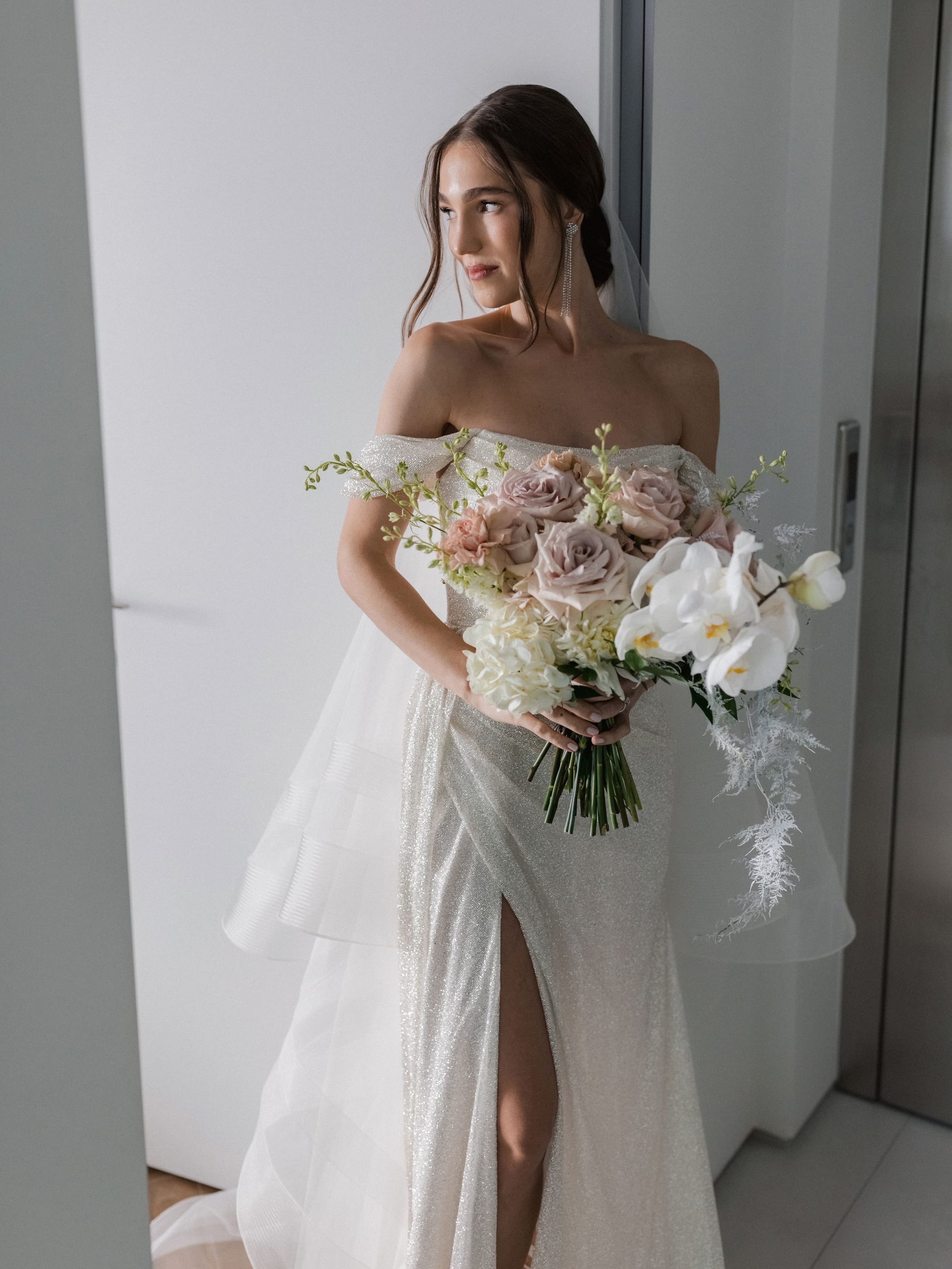 A bride in a shimmering, off-the-shoulder gown with a thigh-high slit stands beside a pale wall, glancing away over her shoulder while holding a cascading bouquet of dusky mauve garden roses, white orchids, cream hydrangea and trailing silver-tipped foliage. This is a wedding bouquet photographed at Amphi Residence in Tria Monastiria, Crete, Greece. Soft, clean daylight and a calm, minimal backdrop give the scene a serene, elegant mood.