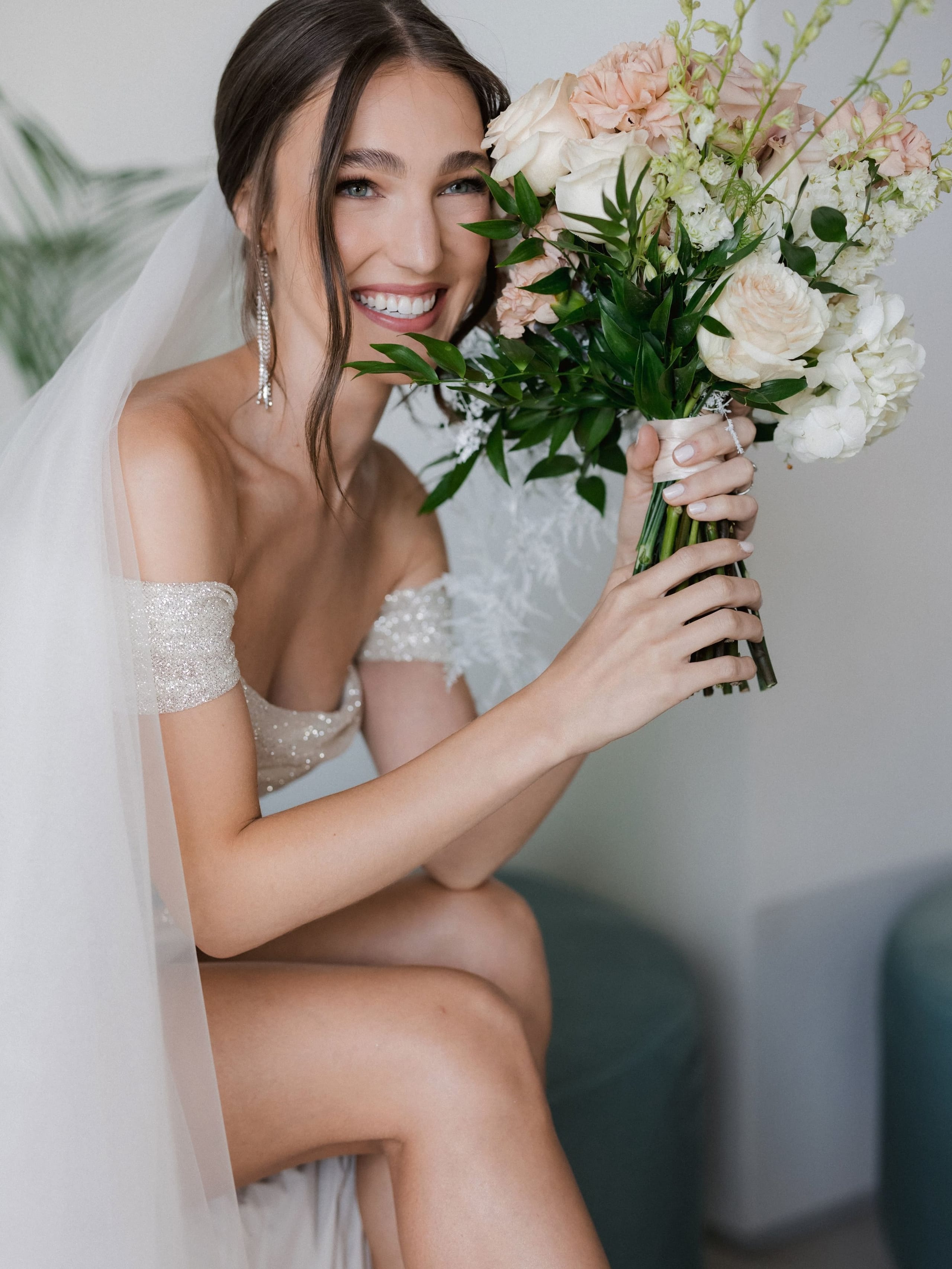 A smiling bride in an off-the-shoulder sequined gown and a sheer veil sits on a teal cushioned seat, holding a bridal bouquet of blush garden roses, white hydrangeas, and white stock, with a few delphinium stems and lush green ruscus leaves framing her face. This wedding flower arrangement was created in Greece for a bride at Amphi Residence in Tria Monastiria, Crete, and the soft, neutral palette glows against the pale walls in natural light.
