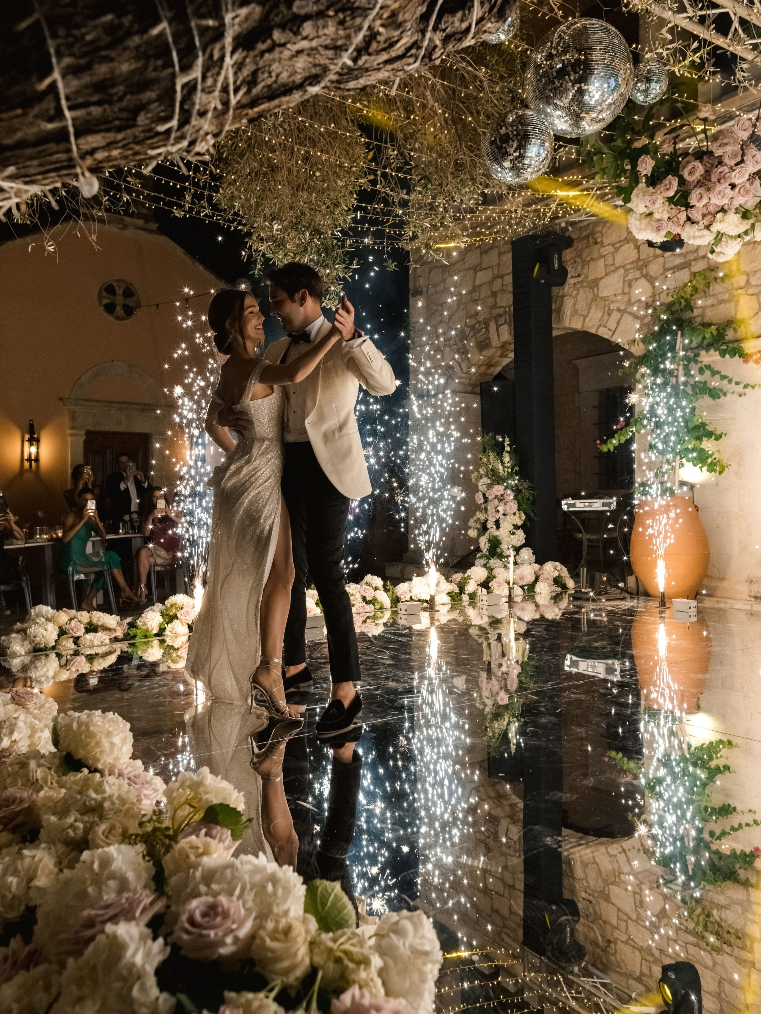 A bride in a sparkling, high-slit gown and a groom in a cream dinner jacket and black bow tie dance under a canopy of disco balls and olive branches, with fountains of sparks rising around them on a glossy reflective floor. Along the edges, pale hydrangeas, blush garden roses and white stock spill across the floor, mirrored in its surface. This is a wedding at Amphi Residence in Tria Monastiria, Crete, Greece.