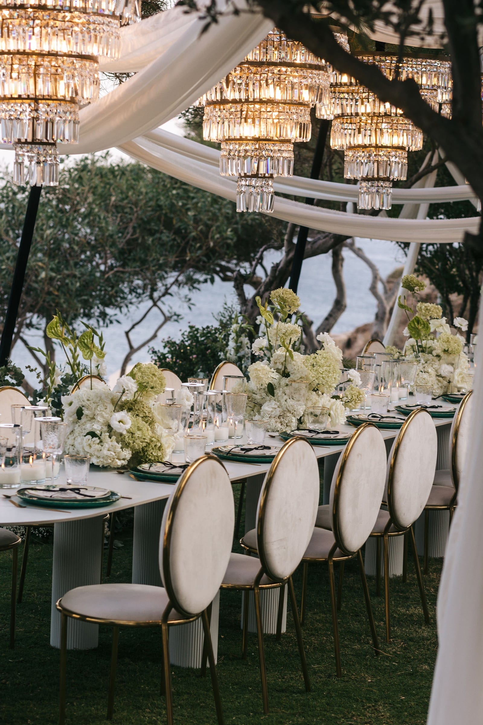 A long wedding dining table under a draped white canopy at Cycladic Gem in Greece, dressed with low centrepieces of white hydrangeas, white roses and green anthurium. Tiered crystal chandeliers hang above, and cream chairs with gold-rimmed backs line the table, which is set with green-rimmed plates and candlelit glassware. Beyond the olive trees, the sea glimmers in the soft evening light.