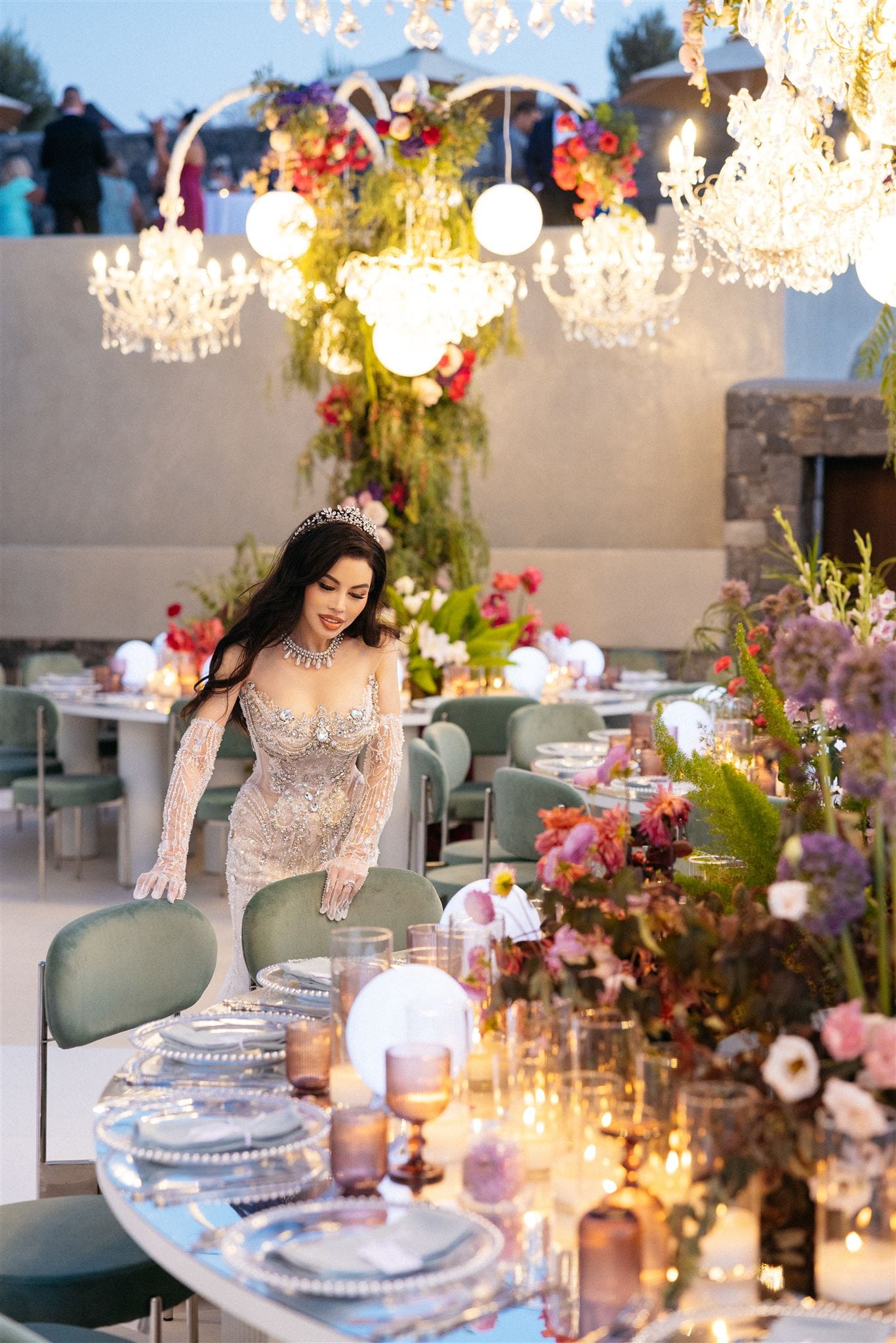 A woman in a heavily beaded, sheer-sleeved gown and a jewelled tiara leans over a green velvet chair at a long wedding table at iIslet in Santorini, Greece. Low tables of mixed flowers in plum, coral and blush, including allium, dahlias and gladioli, fill the foreground, while tall floral installations and crystal chandeliers glow above the room. Pillar candles and frosted glass globes add warm light to the soft, neutral setting.