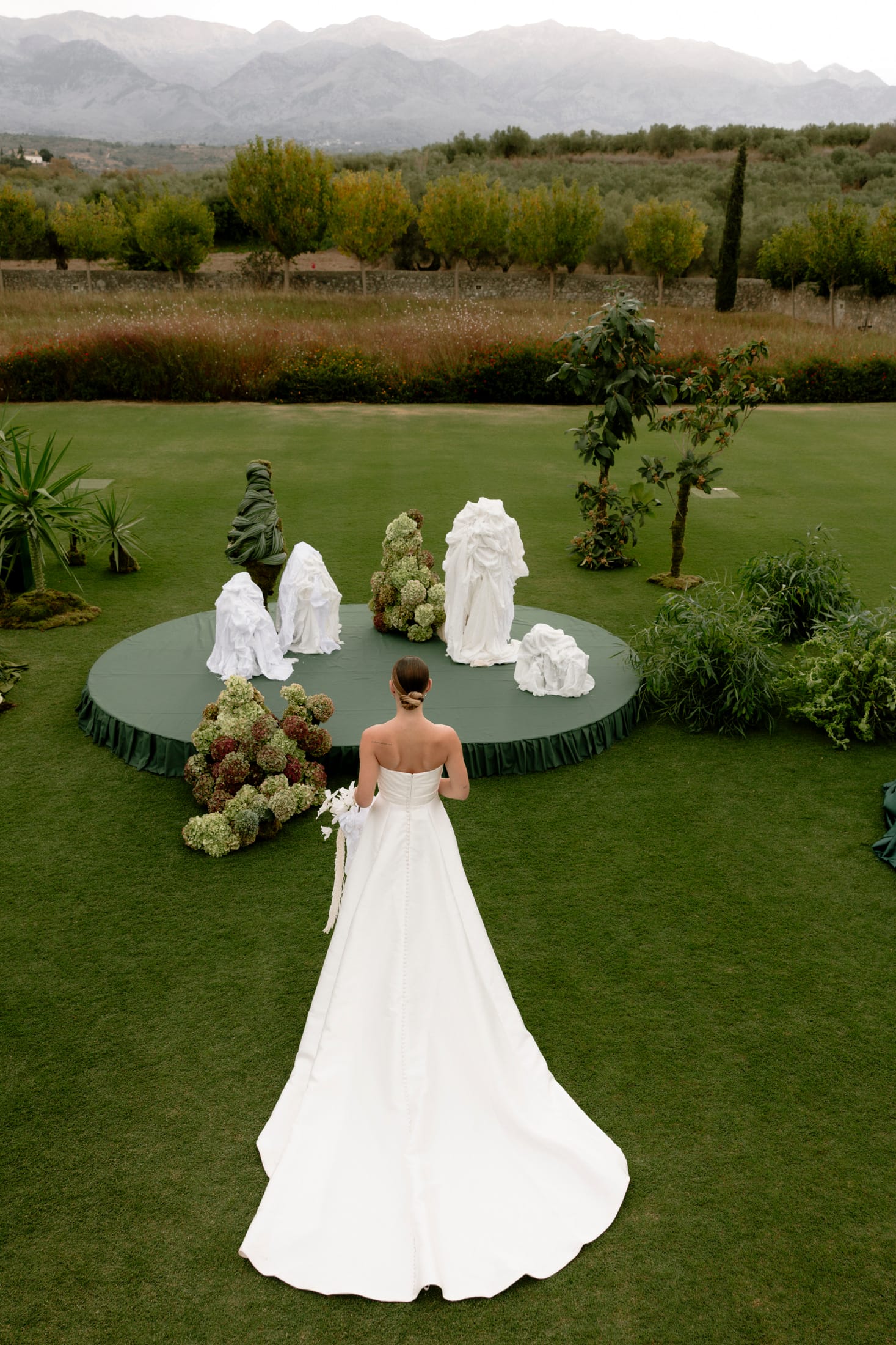 A bride in a strapless white satin gown with covered buttons down her back walks across a sweep of green lawn toward a round dark-green platform. On it stand tall sculptural arrangements of white draped fabric and clusters of green-and-burgundy hydrangeas, with a spiky green foliage piece and two young trees beside them. This wedding flower installation at Roxani Estate in Greece has a calm, garden-like mood, shot from above.