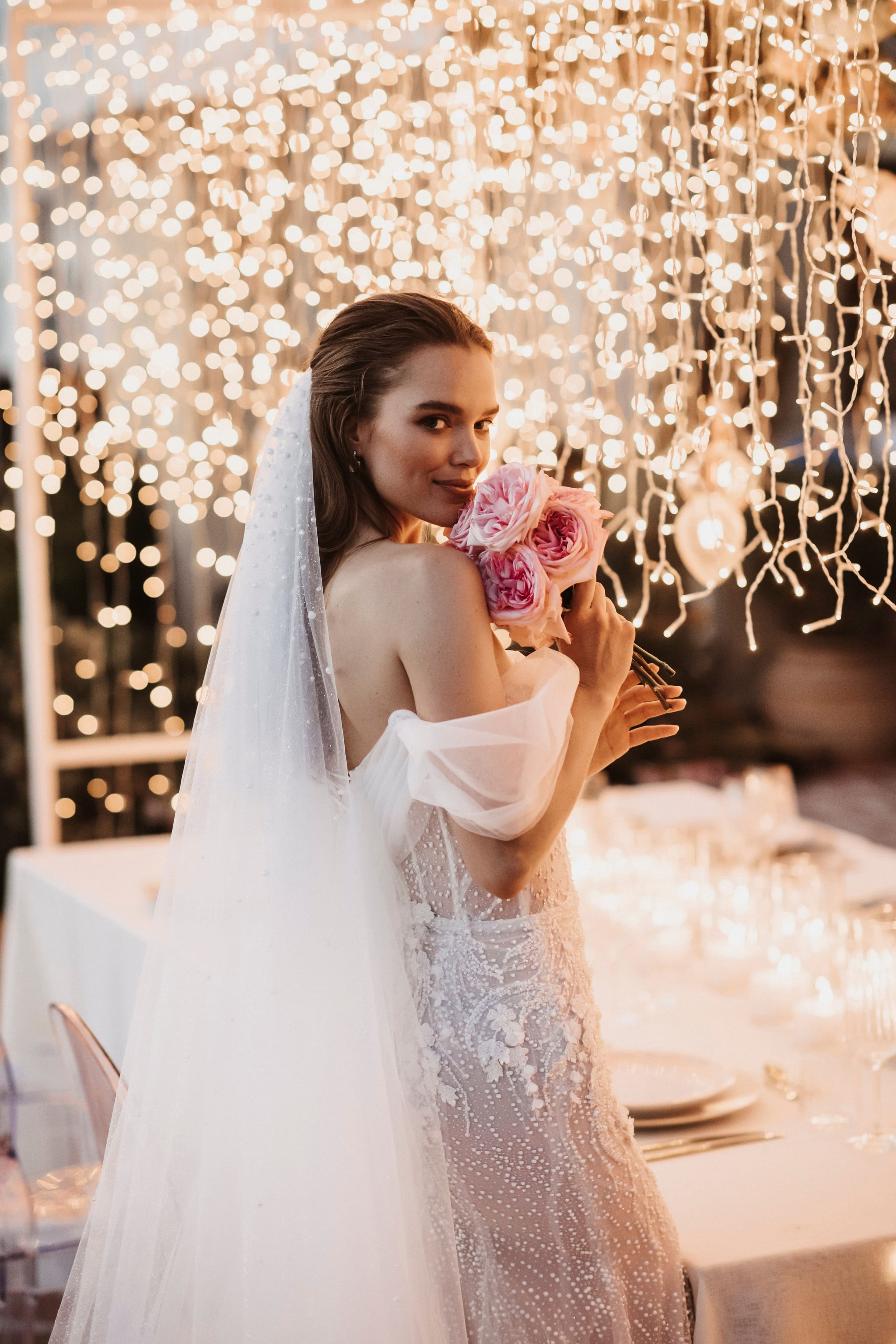 A bride with her dark hair swept back glances over her shoulder, holding a small bouquet of pink garden roses against her shoulder. She wears an off-the-shoulder gown with sheer sleeves, pearl-dotted tulle and a veil edged with pearls. Behind her a curtain of warm fairy lights cascades from above, and a long table set with glassware and white linen glows beside her. This is a wedding flower image at Pandora Suites Hotel in Chania, Crete, Greece.