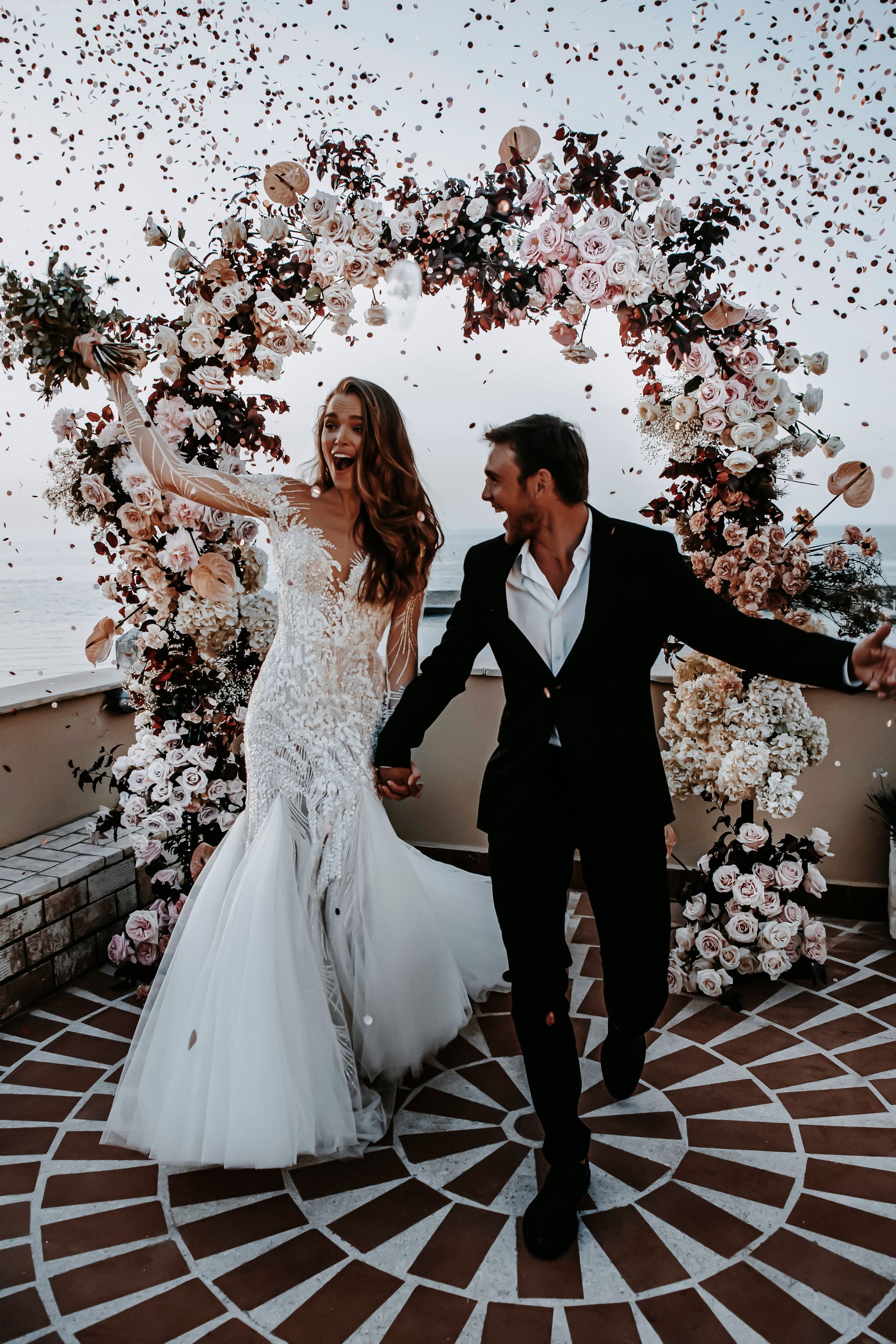 A joyful bride and groom laugh as they walk hand in hand through a shower of confetti beneath a tall floral arch of blush garden roses, cream hydrangeas and deep burgundy foliage, a wedding flower installation at Pandora Suites in Greece. She wears a fitted lace gown with sheer illusion sleeves and a flowing tulle skirt, while he wears a black suit with an open-collared white shirt. Behind them the terrace opens onto a calm sea under a pale sky.