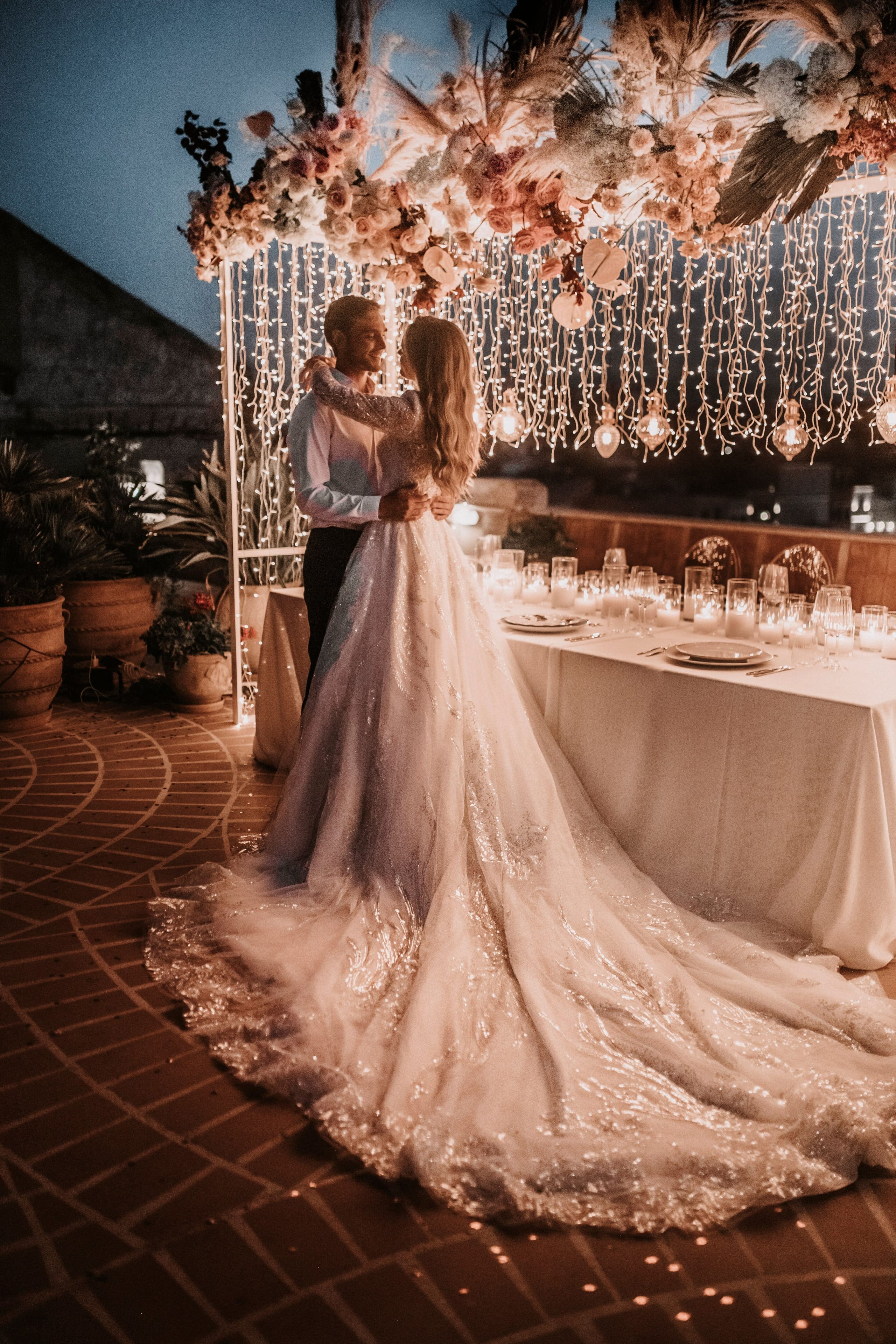 A bride in a long, sparkling gown with a sweeping train and a man in a white shirt and dark trousers embrace under a canopy of pink and cream roses, dried pampas and pale blooms, hung with strings of fairy lights and bulb lanterns. Beside them stands a candlelit dining table on a terrace at Pandora Suites Hotel in Chania, Crete, Greece, set for a wedding dinner. Warm lamplight and terracotta tiles glow against the deep blue evening sky.