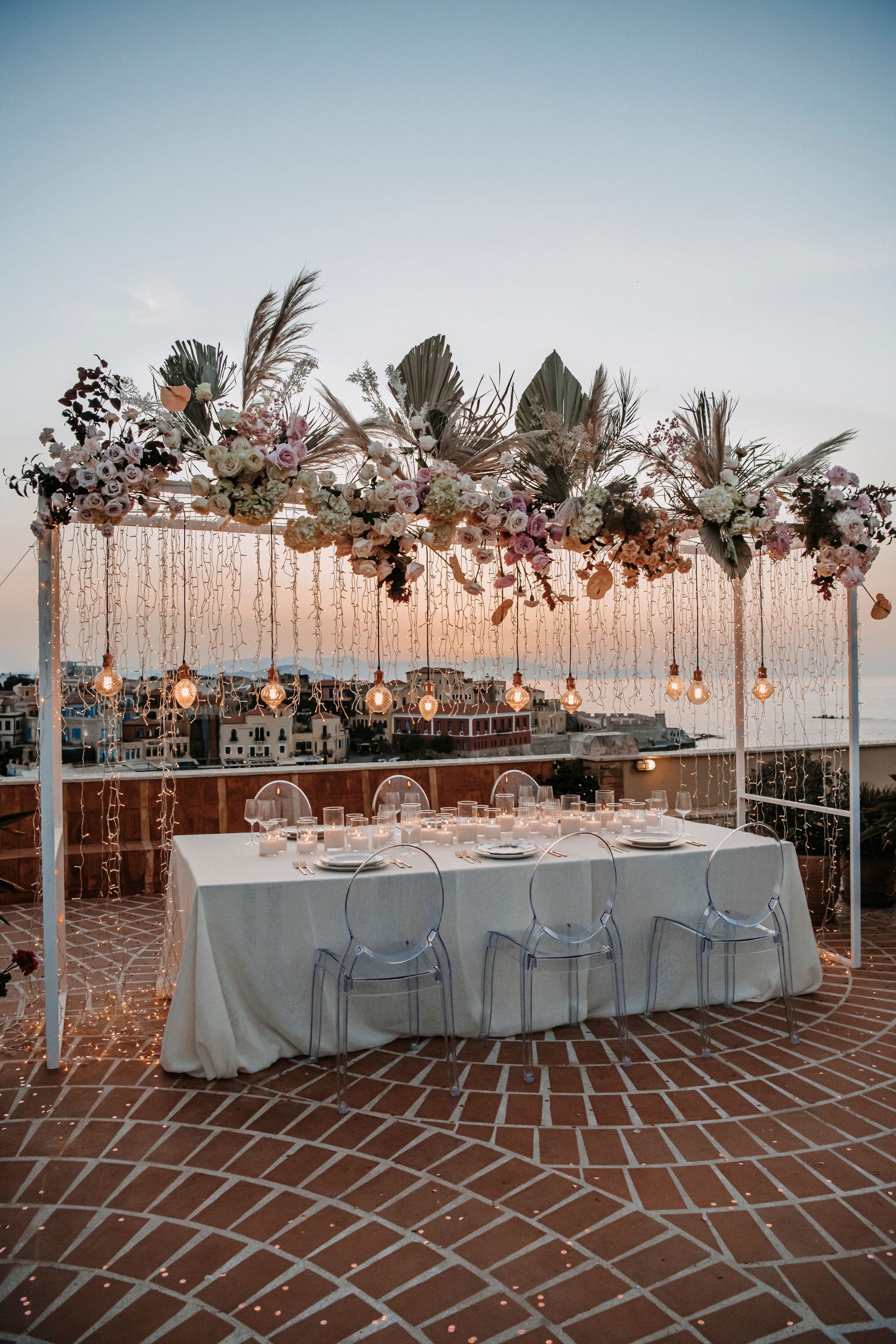 A long white-clothed table set for dinner on a rooftop terrace at Pandora Suites Hotel in Chania, Crete, Greece, with clear acrylic chairs and candles along its length. Overhead, a white frame is draped with fairy lights and hung with a lush floral canopy of pale roses, white hydrangeas, dusky pink blooms and dried pampas and palm fronds. Warm bulbs glow beneath it as the sky fades to dusk above the town's rooftops and sea.