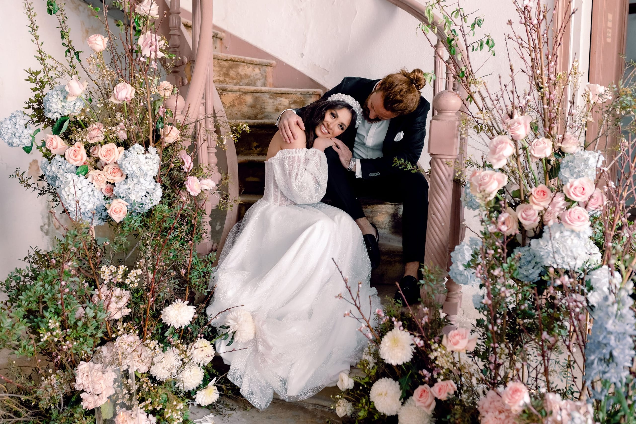 A bride in a sparkly white gown with puffed sheer sleeves and a pearl headband rests her cheek against the shoulder of a groom in a dark suit, who leans in beside her on a weathered, rose-painted spiral staircase. Both are smiling softly. The staircase is framed on each side by lush arrangements of pale pink garden roses, blush hydrangeas, white chrysanthemums, and flowering branches, all part of the wedding flower styling in Greece.