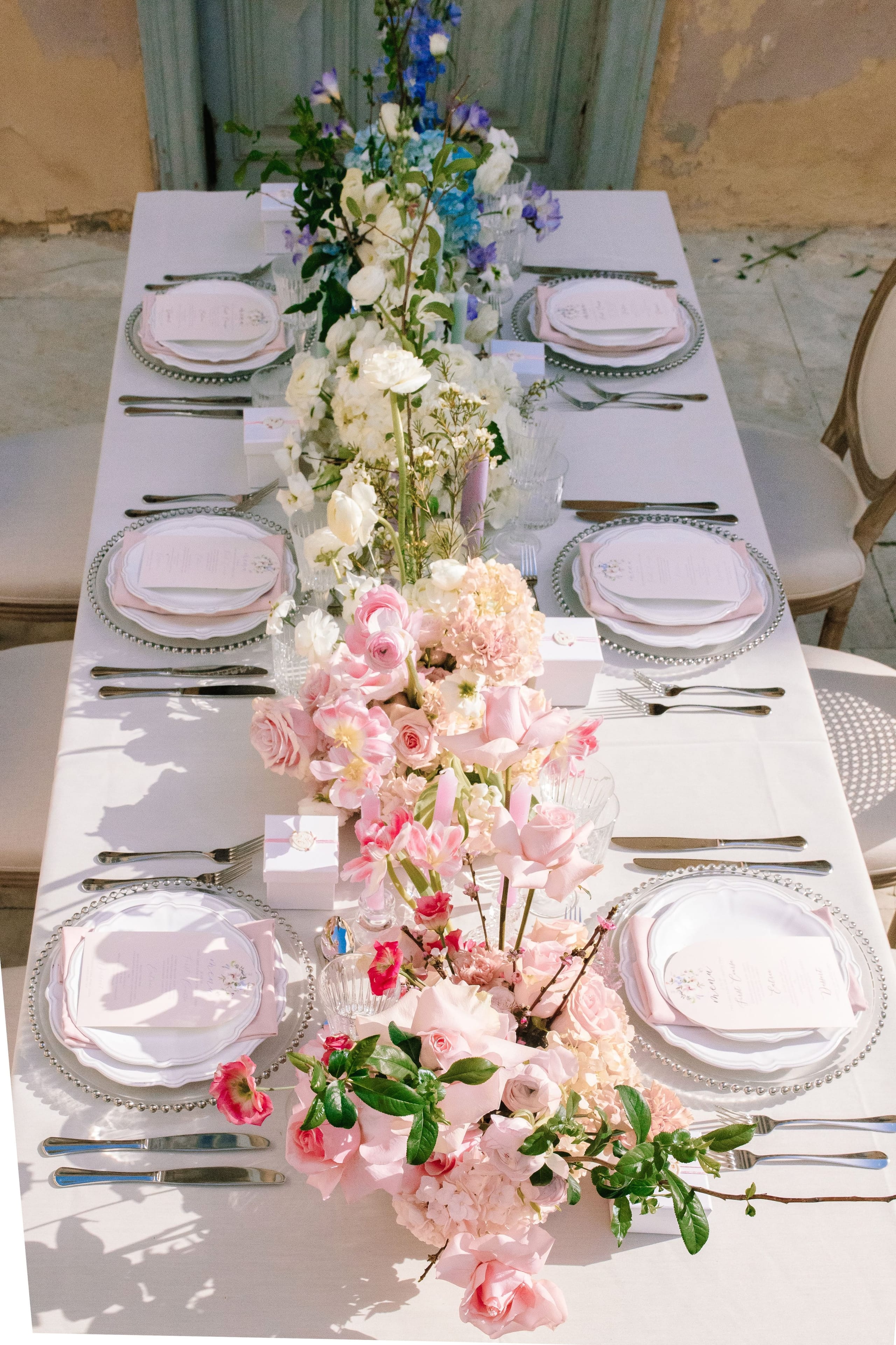 A long white-draped wedding table in Greece, set with beaded silver charger plates, pale pink napkins and menu cards, and a central runway of pink garden roses, blush hydrangeas, pink tulips and ranunculus, with white blooms and sprigs of blossom threaded through. Soft sunlight falls across the linen, and the whole setting feels delicate and romantic.