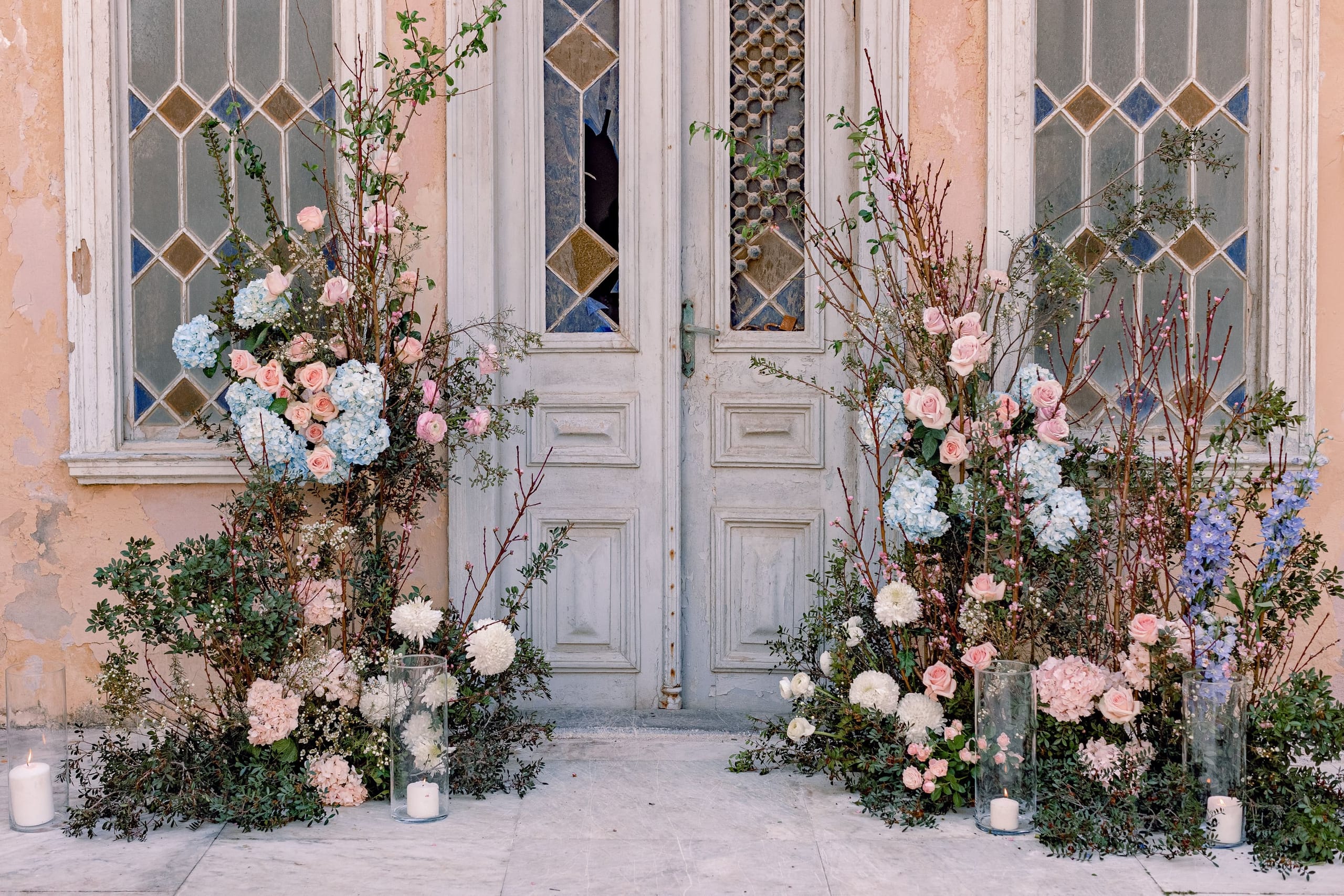 Two tall floral installations frame a pair of weathered grey wooden doors with leaded diamond-pane glass, set against a faded peach wall in Greece. Each side is filled with pale pink garden roses, blue hydrangeas, white dahlias, bare branches with blush blossoms and trailing greenery, with clear glass hurricane vases holding lit candles at the base. It is wedding flowers in Greece, styled as a soft, romantic entrance for the ceremony.