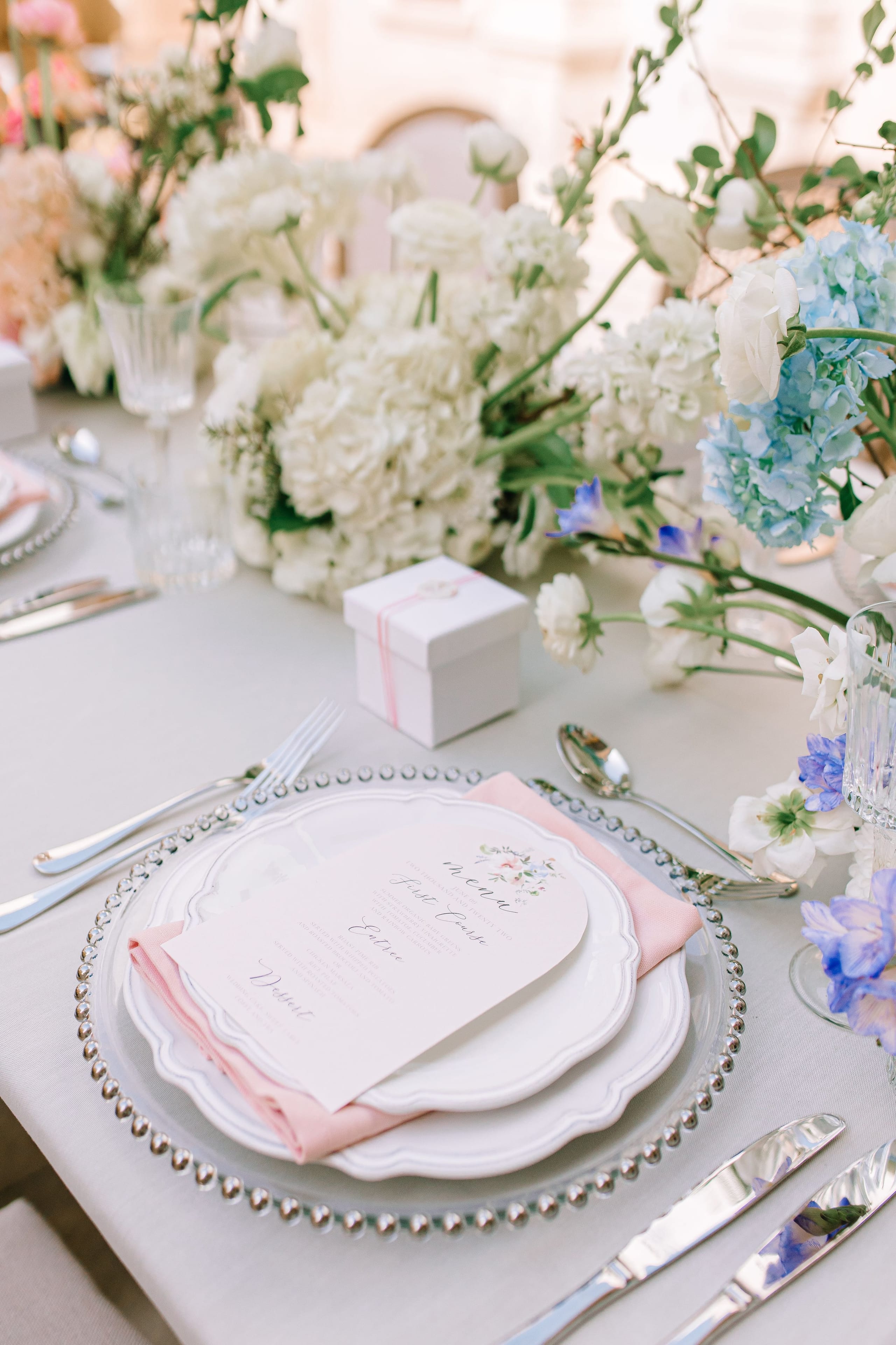 A wedding table set for Alysia and Fraunk in Greece, with a pale pink menu card resting on a beaded glass charger beside silver cutlery and a pink napkin. Behind it, a centrepiece of creamy white hydrangeas and ranunculus is mixed with pale blue hydrangeas and sprigs of purple delphinium. A small white gift box tied with a pink ribbon sits at the centre, and the soft, airy palette feels delicate and romantic.