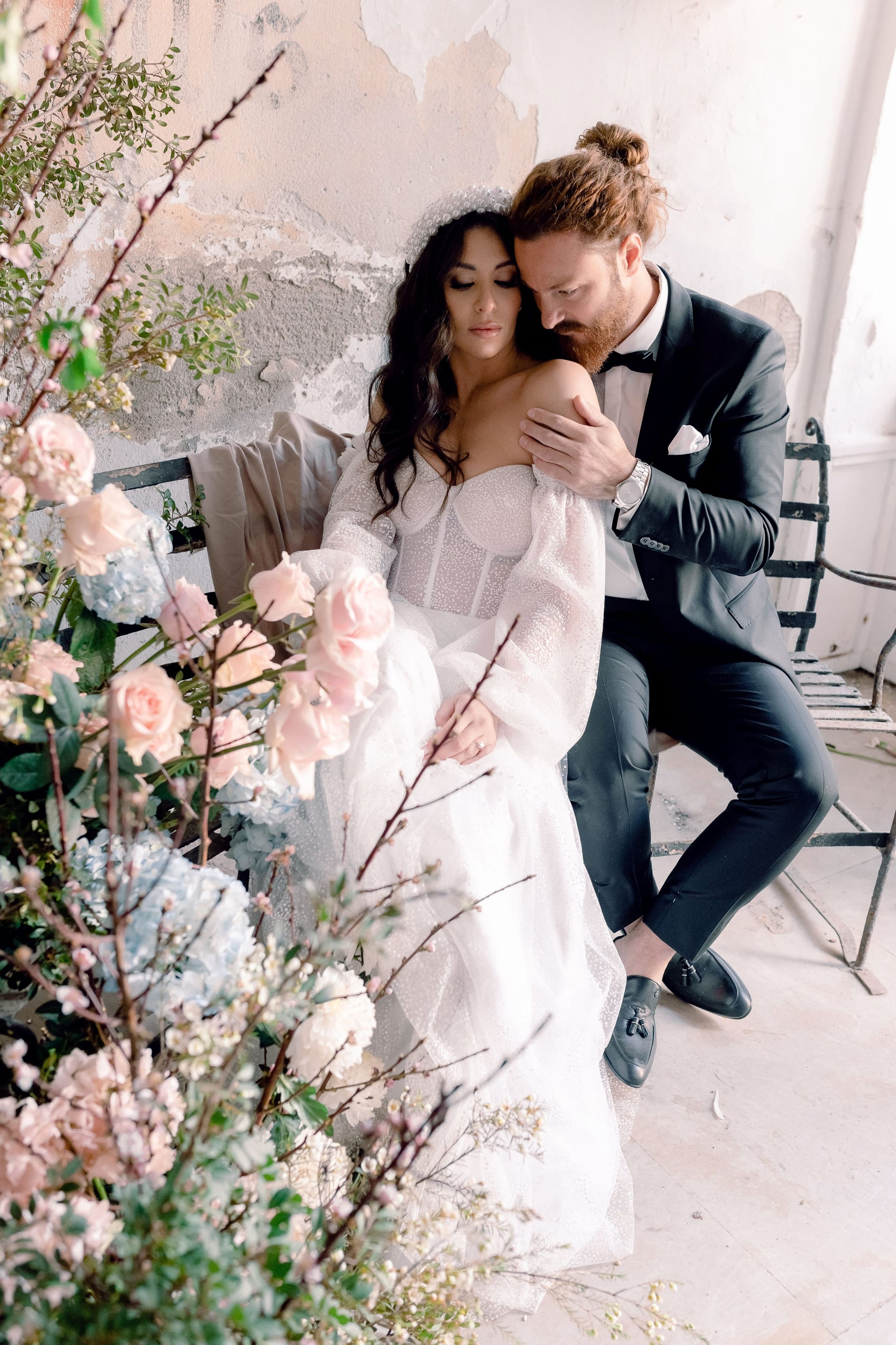 A bride in a sheer, dotted-tulle gown with puffed sleeves and a corset bodice sits on a weathered iron bench, while a groom in a dark tuxedo leans in to rest his hand on her shoulder. In the foreground, a lush arrangement of pale pink garden roses, blue hydrangeas and bare blossom branches frames the couple against a crumbling plaster wall. This wedding flower styling in Greece has a soft, airy palette of blush, pale blue and cream.