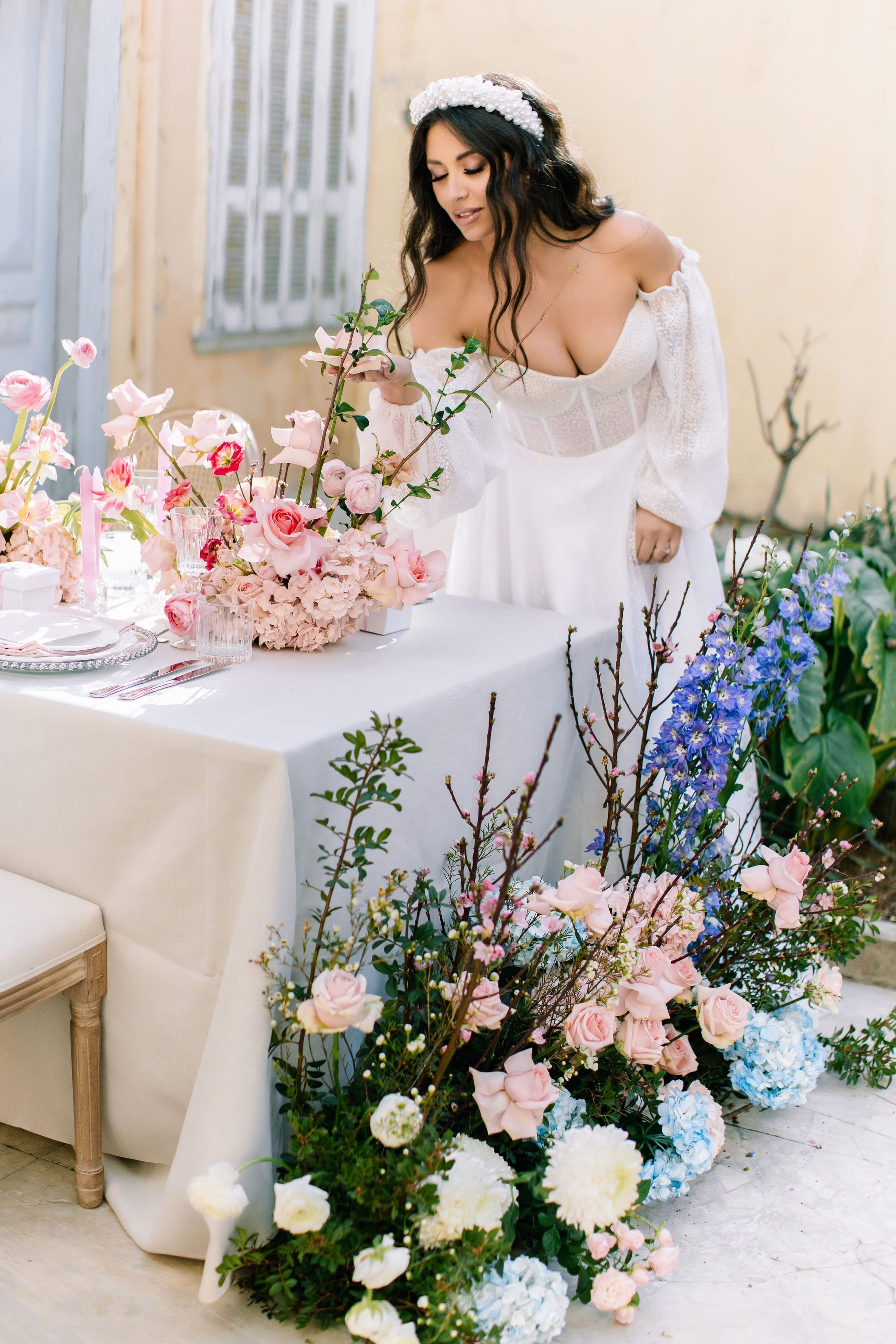 A lush, low floral installation spills along the base of a draped white sweetheart table at the Alysia & Fraunk wedding in Greece. Blush garden roses, pale pink hydrangeas, white ranunculus and a white dahlia are tucked among blue hydrangeas, tall violet-blue delphinium spires, and bare pink-tipped branches and sprigs of green foliage. The soft pastel palette glows in bright daylight on a pale marble terrace.
