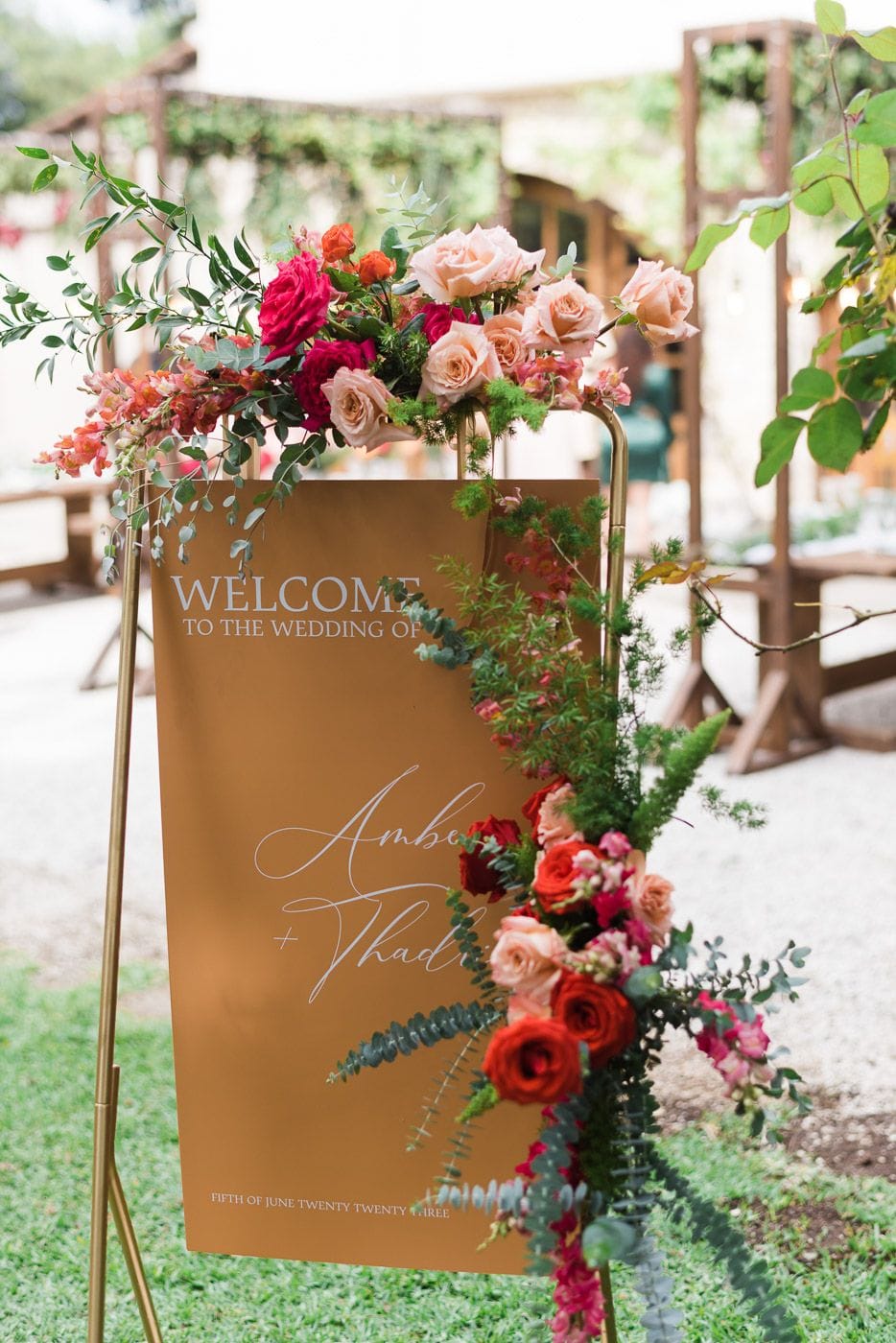 A tall gold easel welcome sign reading "Welcome to the wedding of" in white script, with the names Amber and Thadrick, stands on grass at Dourakis Winery in Alikampos, Crete, Greece. Cascading down its right side and across the top are wedding flowers in blush peach and coral garden roses, deep red roses, orange ranunculus, and pink snapdragons, all woven through trailing eucalyptus and feathery green foliage. Soft, blurred wooden arbours and gravel in the background suggest a warm, relaxed garden setting.