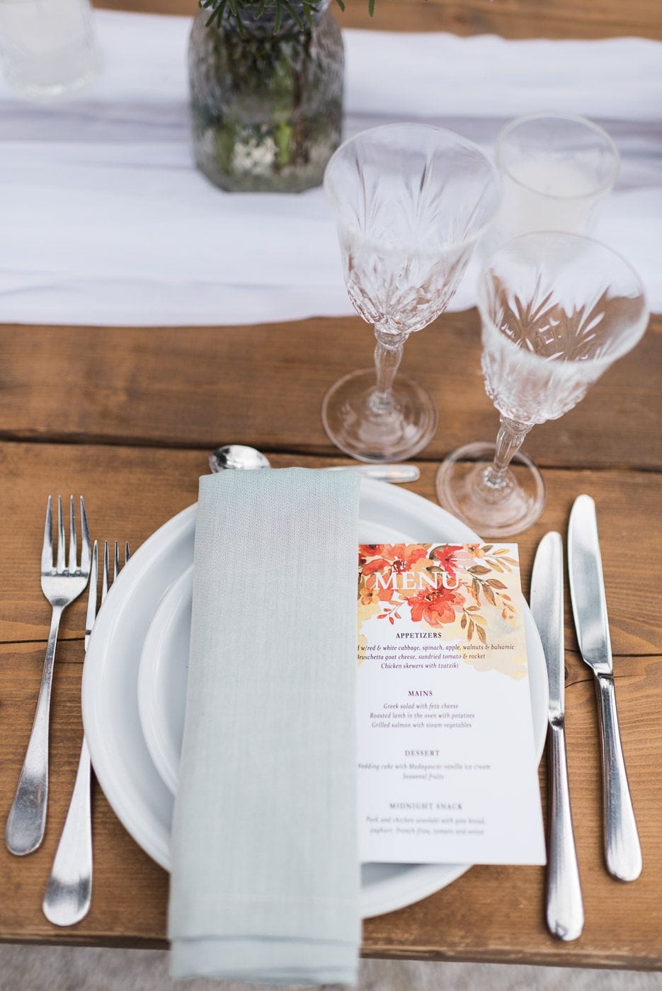 A place setting on a rustic wooden table at Dourakis Winery in Alikampos, Crete, Greece, for a wedding. A pale sage-grey linen napkin sits on a white plate beside silver cutlery and cut-glass wine glasses, with a small glass jar of greenery and a white table runner behind. A printed menu card with orange and red watercolour florals rests against the plate.