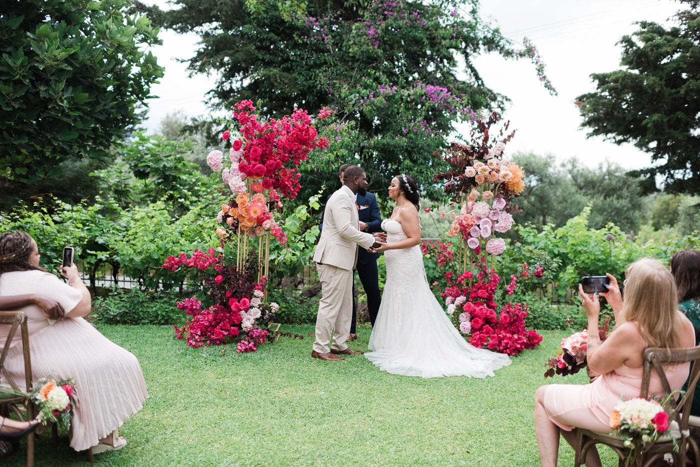 A bride in a fitted white lace gown with a long train and a pearl hair accessory stands facing her groom in a light stone-coloured suit on a lawn at Dourakis Winery in Alikampos, Crete, Greece, holding his hands. Two tall wedding arches of magenta bougainvillea, hot pink roses, peach garden roses and deep burgundy foliage frame them, with a pale blush rose cluster on the right. Guests in blush pink dresses sit on rustic wooden chairs at the aisle's edge, one raising a phone to capture the moment, under leafy green trees and vines.