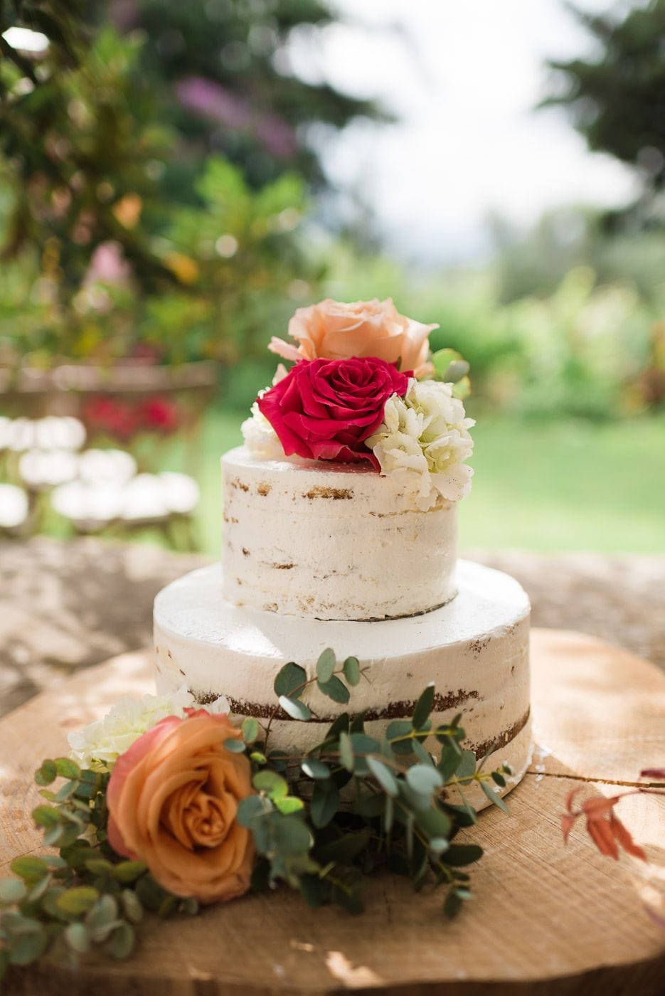 A two-tier naked wedding cake, its pale buttercream showing the sponge in rustic streaks, sits on a round wooden slice outdoors. Its top is crowned with a deep pink garden rose, a soft apricot rose and white hydrangea blooms, while more apricot rose and sprigs of silvery eucalyptus trail across the wood in the foreground. The garden behind is softly blurred in green, lit by warm sunshine, at Dourakis Winery in Alikampos, Crete, Greece, a wedding cake for Amber and Thadrick.