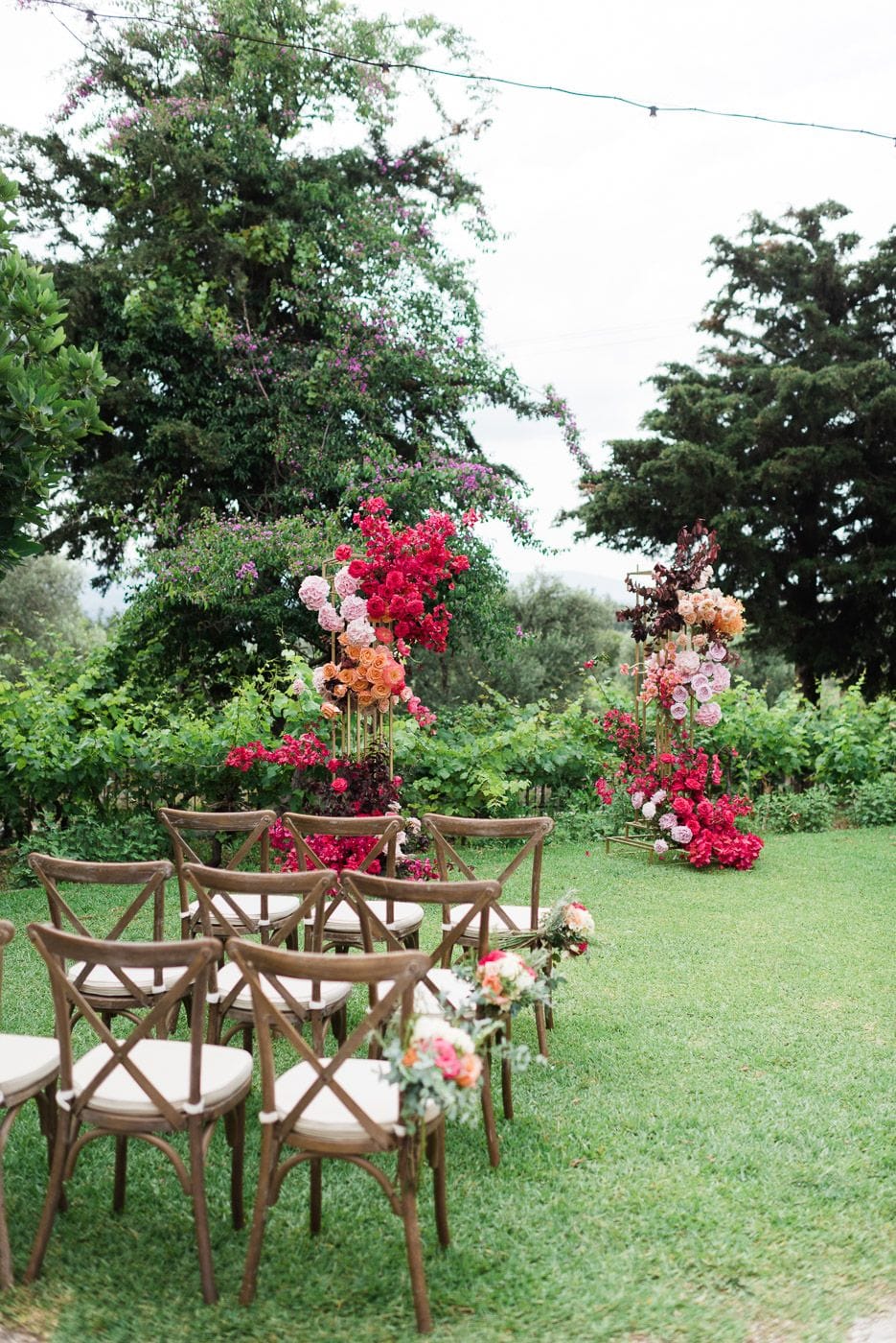 Rows of wooden cross-back chairs with cream cushions sit on a lush green lawn, leading toward two tall gold stands wrapped in cascades of flowers. The left stand blooms with hot pink bougainvillea, pale pink and peach roses, and deep burgundy foliage, while the right is draped in blush and coral roses. Set among vineyard greenery and tall trees at Dourakis Winery in Alikampos, Crete, Greece, this is a wedding ceremony aisle styled with vibrant, romantic floral arches.
