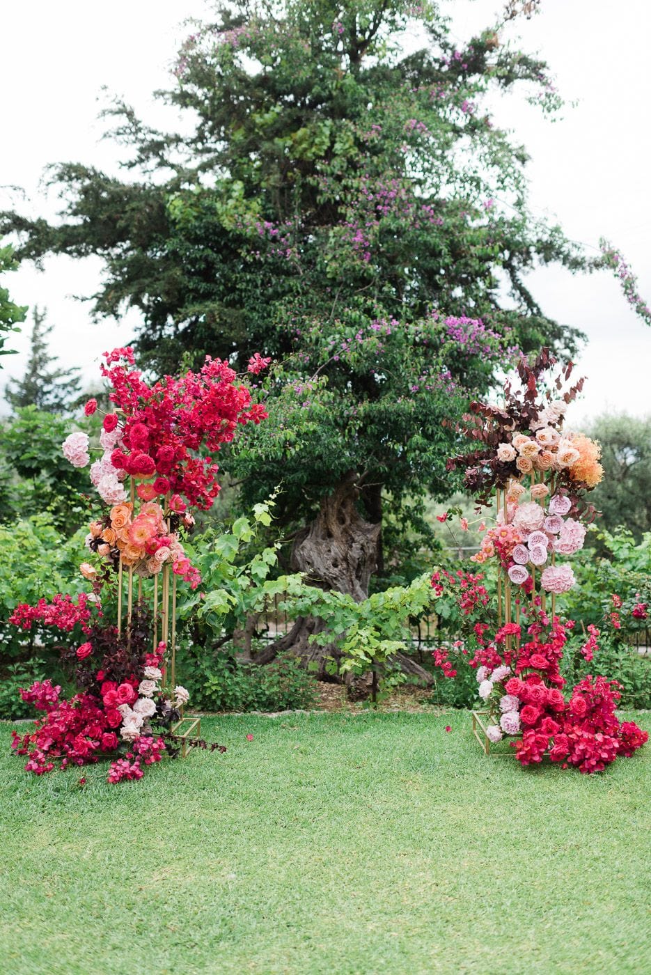 Two tall gold metal stands on a lawn frame an aisle, each one bursting with wedding flowers in a gradient from hot pink and crimson at the bottom to soft blush and apricot at the top. The left stand is full of bougainvillea and orange garden roses, the right one of pale pink roses, peach blooms and dark burgundy foliage, all set at Dourakis Winery in Alikampos, Crete, Greece. Behind them a large old tree covered in purple bougainvillea makes a lush green backdrop under a soft, overcast sky.