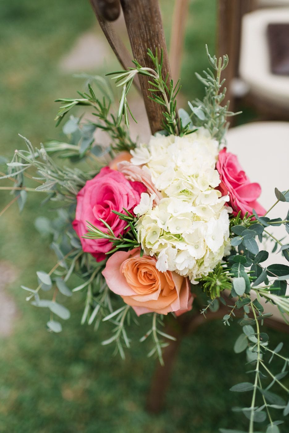 A small arrangement of cream hydrangea, two hot pink garden roses and a peach-apricot rose tied with trailing eucalyptus and sprigs of rosemary, hung from the wooden back of a chair on lawn. This is a wedding floral decoration for the aisle at Dourakis Winery in Alikampos, Crete, Greece, with the soft green grass and blurred chairs behind it.
