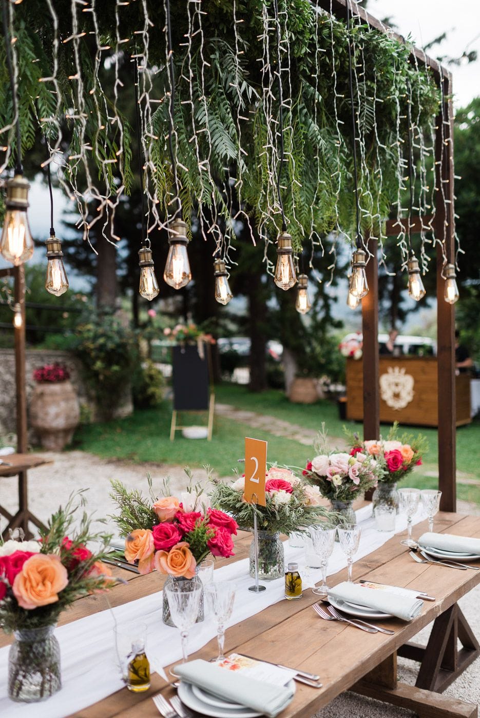 A long rustic wooden wedding table in a garden at Dourakis Winery in Alikampos, Crete, Greece, set with a white runner and small glass vases of hot pink and peach roses and sprigs of greenery. Overhead, a wooden pergola hung with feathery green foliage and strings of Edison bulbs and fairy lights glows warmly, while an orange table number "2" stands among the vases. Behind the table, lawn and a wooden bar are softly blurred in the evening light.