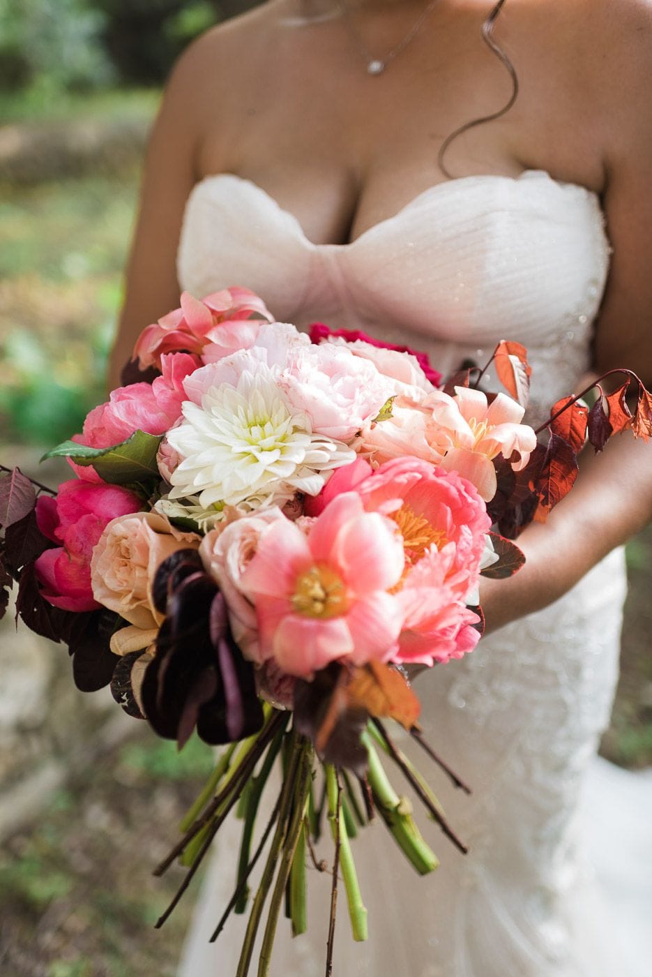 A bride in a strapless white gown holds a loose, cascading bridal bouquet of pink and coral peonies, blush garden roses, a white dahlia and deep pink ranunculus, with dark plum leaves and rust-coloured autumn foliage trailing from the stems. The bouquet, a wedding flower arrangement photographed at Dourakis Winery in Alikampos, Crete, Greece, is softly lit against a blurred green background.