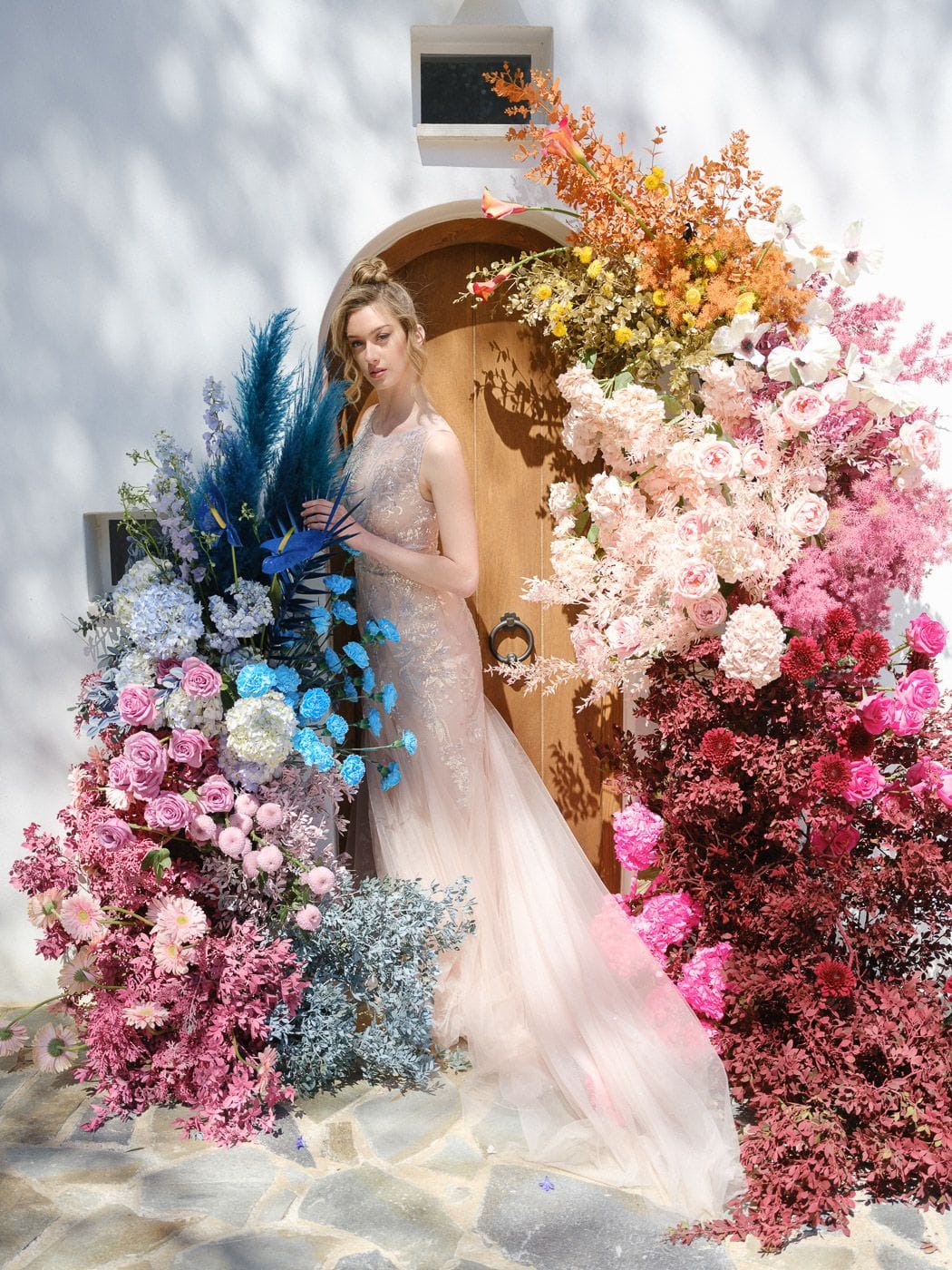 A bride in a blush tulle gown with delicate embroidery stands in front of an arched wooden door, framed by a towering floral installation that shifts from deep blue and lavender on the left to pink, peach and burgundy on the right. Blue pampas plumes, delphinium and carnations mingle with white hydrangea and pink roses, while orange and gold foliage arcs overhead. These wedding flowers, set against whitewashed walls at Markakis Estate in Emprosneros, Crete, Greece, make a vivid, sunlit display.