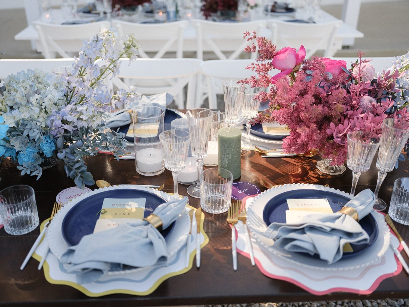 A long dark wooden wedding table set outdoors at Markakis Estate in Emprosneros, Crete, Greece, with two place settings in front. On the left, pale blue hydrangeas and lilac delphinium spill from a low arrangement with eucalyptus, and on the right, hot pink roses and feathery pink astilbe create a bolder centrepiece. Navy plates, pale blue linen napkins, glass tumblers and candles in clear holders sit between the blooms, with white chairs softly blurred behind.