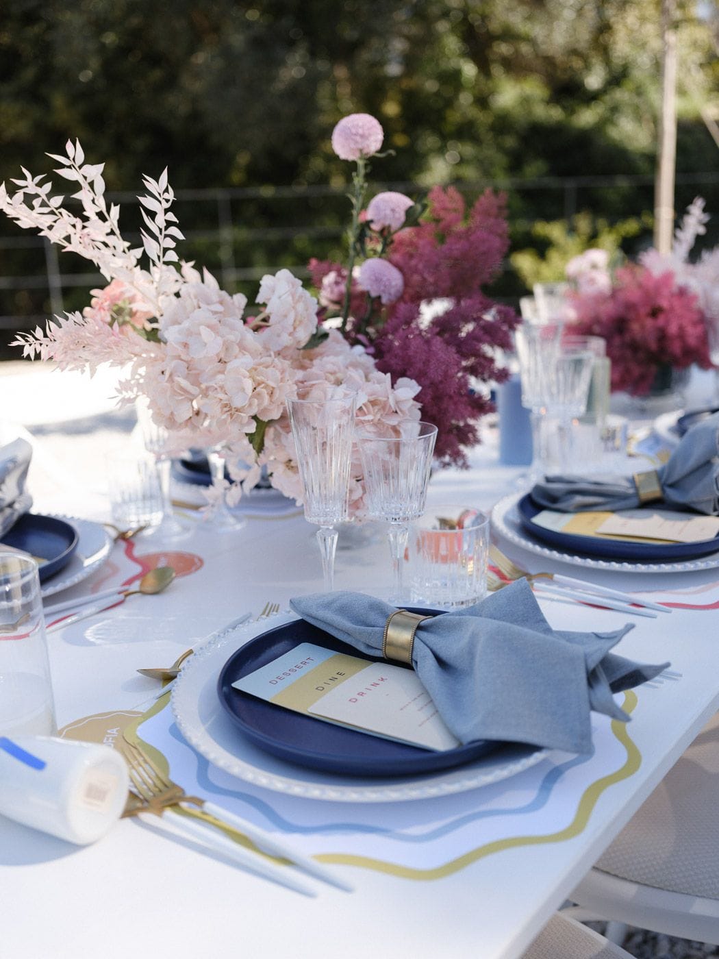 A long outdoor table set for a wedding meal at Markakis Estate in Emprosneros, Crete, Greece, with navy-blue plates rimmed in white, dusky blue linen napkins held by gold rings, and clear crystal glasses. Low arrangements of blush hydrangeas, pale pink feathery plumes and deep magenta astilbe rise beside pink dahlia-like blooms, with sprays of wispy foliage reaching up. Soft afternoon light falls across the white tablecloth and wavy gold-and-blue placemats, with dark green trees blurred behind.