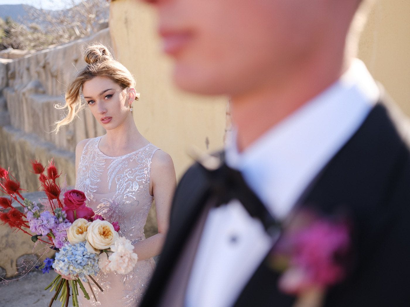 A bride with her hair in a loose bun and soft tendrils, wearing a sleeveless gown with fine beaded lace, glances over her shoulder while holding a bouquet of cream garden roses, hot pink roses, blue hydrangea, lavender stock and red thistle. In the soft-focus foreground a groom in a black suit and white shirt, with a pink bloom on his lapel, is blurred out of focus. This bridal bouquet is by Markakis Estate, a wedding florist in Emprosneros, Crete, Greece, shot against a warm stone wall in bright daylight.