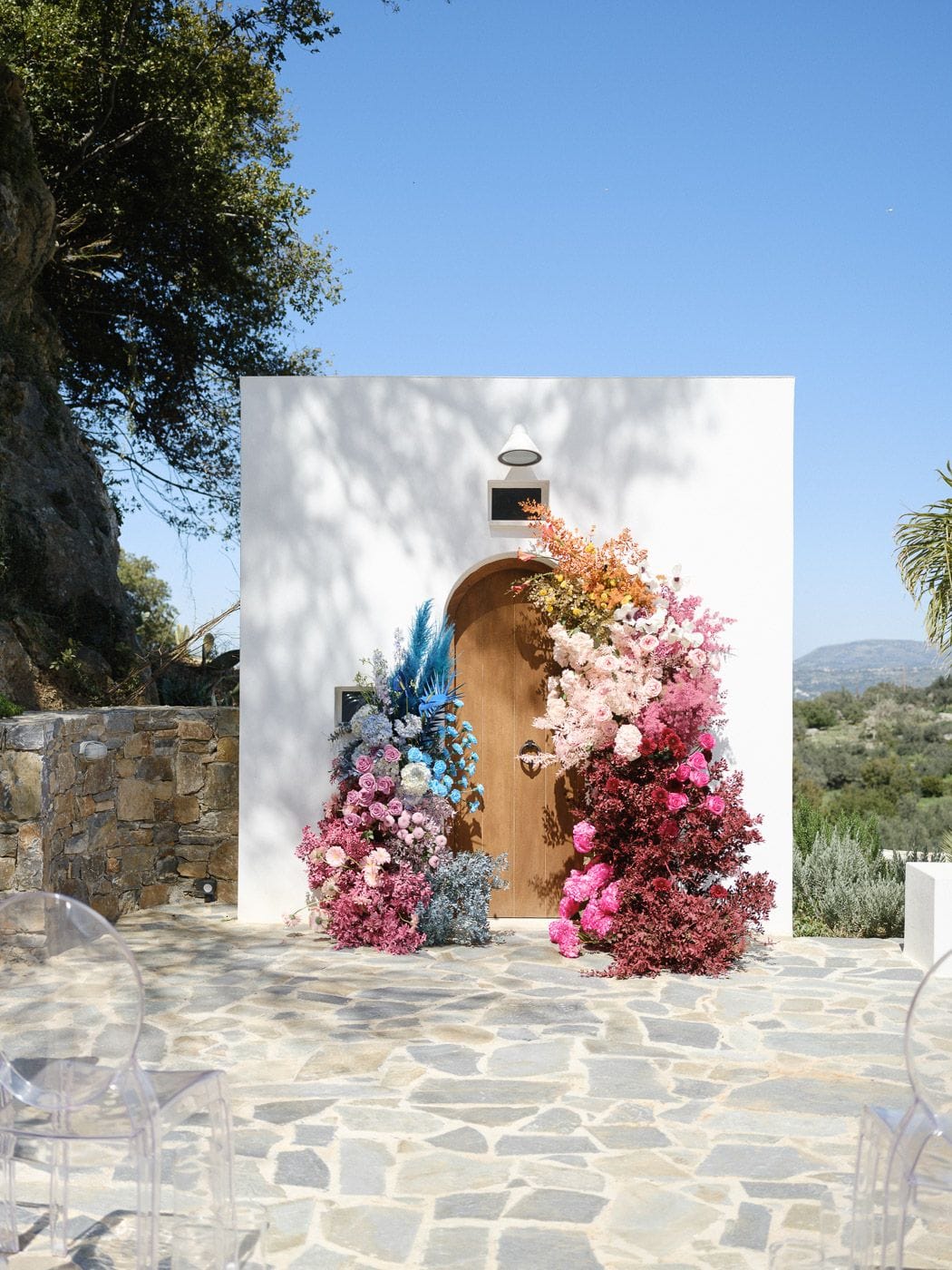 Two flower installations frame an arched wooden door set into a whitewashed wall, creating a wedding ceremony backdrop at Markakis Estate in Emprosneros, Crete, Greece. On the left, blue-tinted pampas grass, lavender-blue flowers and pale pink roses rise from the stone floor, while on the right a gradient climbs from orange and gold through blush pink to deep burgundy, with pink peonies at its base. Clear Ghost chairs sit on the flagstone terrace under a bright blue sky, with hills and olive trees beyond.