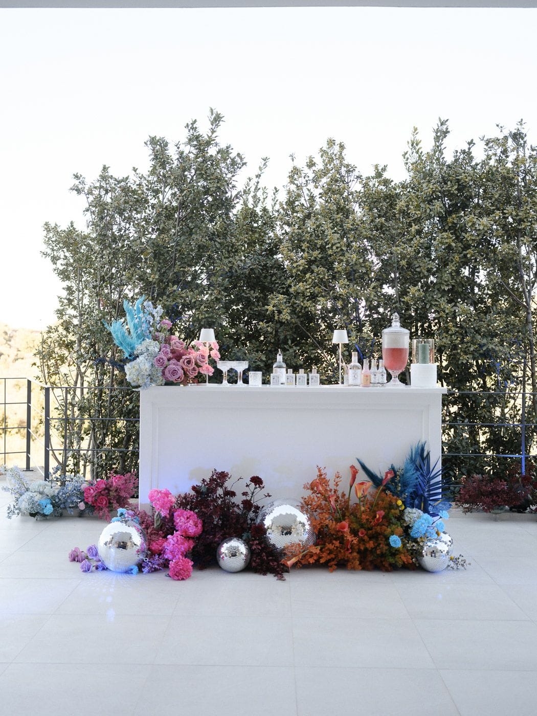 A white bar counter on a pale tiled terrace at Markakis Estate in Emprosneros, Crete, Greece, dressed with wedding flowers for Amillia and Eriks' celebration. Low floral arrangements sweep along its base in hot pink peonies, deep burgundy foliage, orange calla lilies and sky-blue hydrangeas, with large and small mirror disco balls scattered among them. On the counter sit a glass apothecary jar of pink drink, slim lamps and a few bottles, with pink and mauve roses and pale blue hydrangeas at one end, all set against a backdrop of dense green trees under a bright sky.