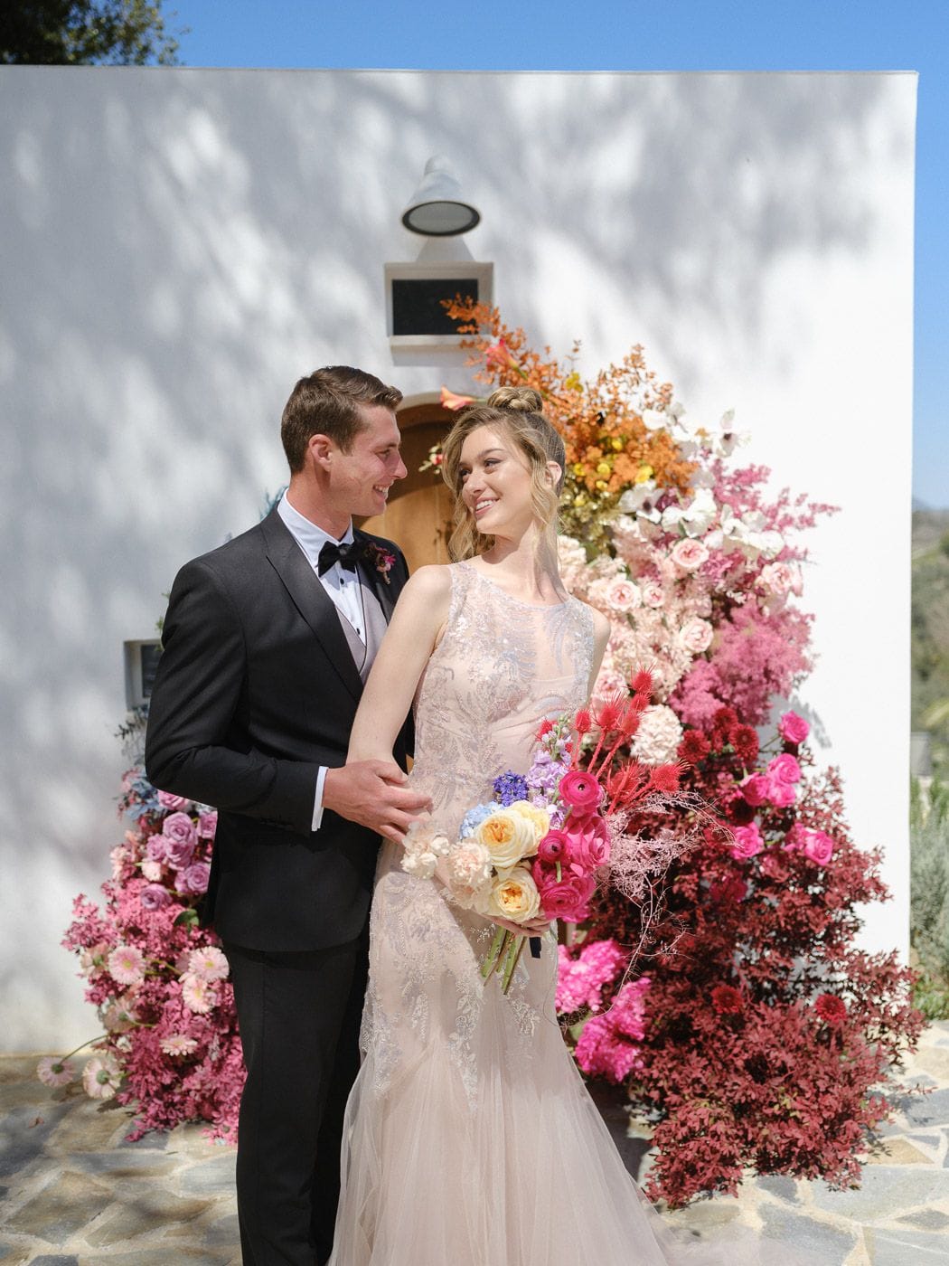 A bride in a pale, lace-embroidered gown and a groom in a black tuxedo with bow tie stand smiling at each other in front of a whitewashed wall, beside a dramatic floral installation of pink and magenta blooms fading to orange and deep red. The bride carries a bouquet of cream roses, pink ranunculus and blue hydrangea, and the scene is wedding flowers at Markakis Estate in Greece.