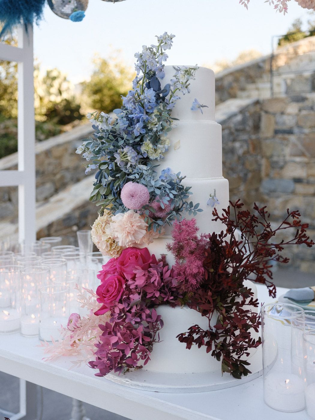 A five-tier white wedding cake draped with a cascade of flowers that shift from pale blue delphinium and eucalyptus at the top, through dusty pink pompoms and cream hydrangea, to hot pink garden roses and magenta blooms at the base, with trailing burgundy foliage on one side. The cake stands on a white table outdoors at Markakis Estate in Emprosneros, Crete, Greece, with rows of glass votive candles and a stone wall behind it in soft evening light.