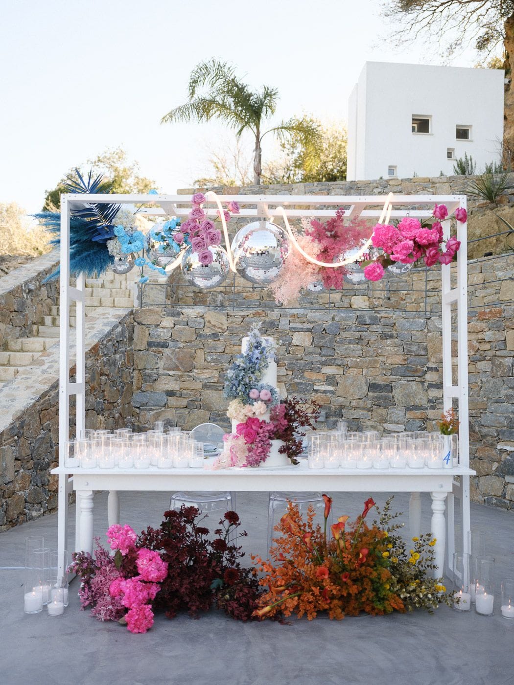 A wedding dessert table set against a stone terrace in Emprosneros, Crete, Greece, at Markakis Estate. Beneath a white frame strung with silver disco balls and draped with pink and blue blooms, a tiered white cake sits decorated with blue delphinium, pale peonies and magenta flowers. Lined with rows of small glowing candles, the table is anchored at its base by lush drifts of hot pink hydrangea, deep burgundy foliage and orange calla lilies.