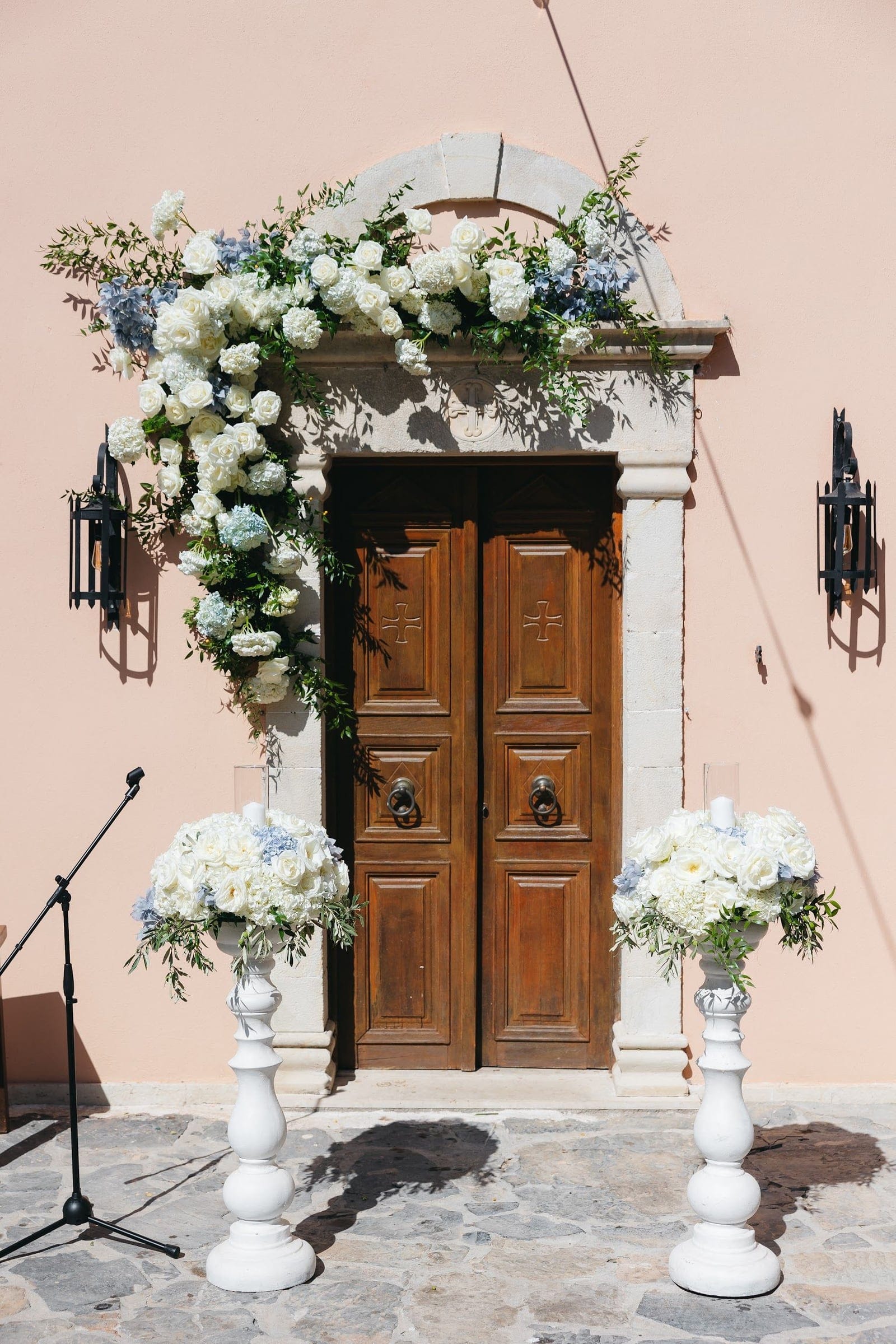 A cascading arrangement of white roses, white hydrangeas and pale blue hydrangeas, threaded with olive foliage, drapes over the stone arch of a pale pink wall, framing a pair of heavy carved wooden doors. Two white pedestal urns, each overflowing with white blooms and greenery, stand on the stone paving before the entrance, with a microphone stand at the left edge. This is a wedding ceremony setting in Crete, Greece, at AgrecoFarms in Adele.