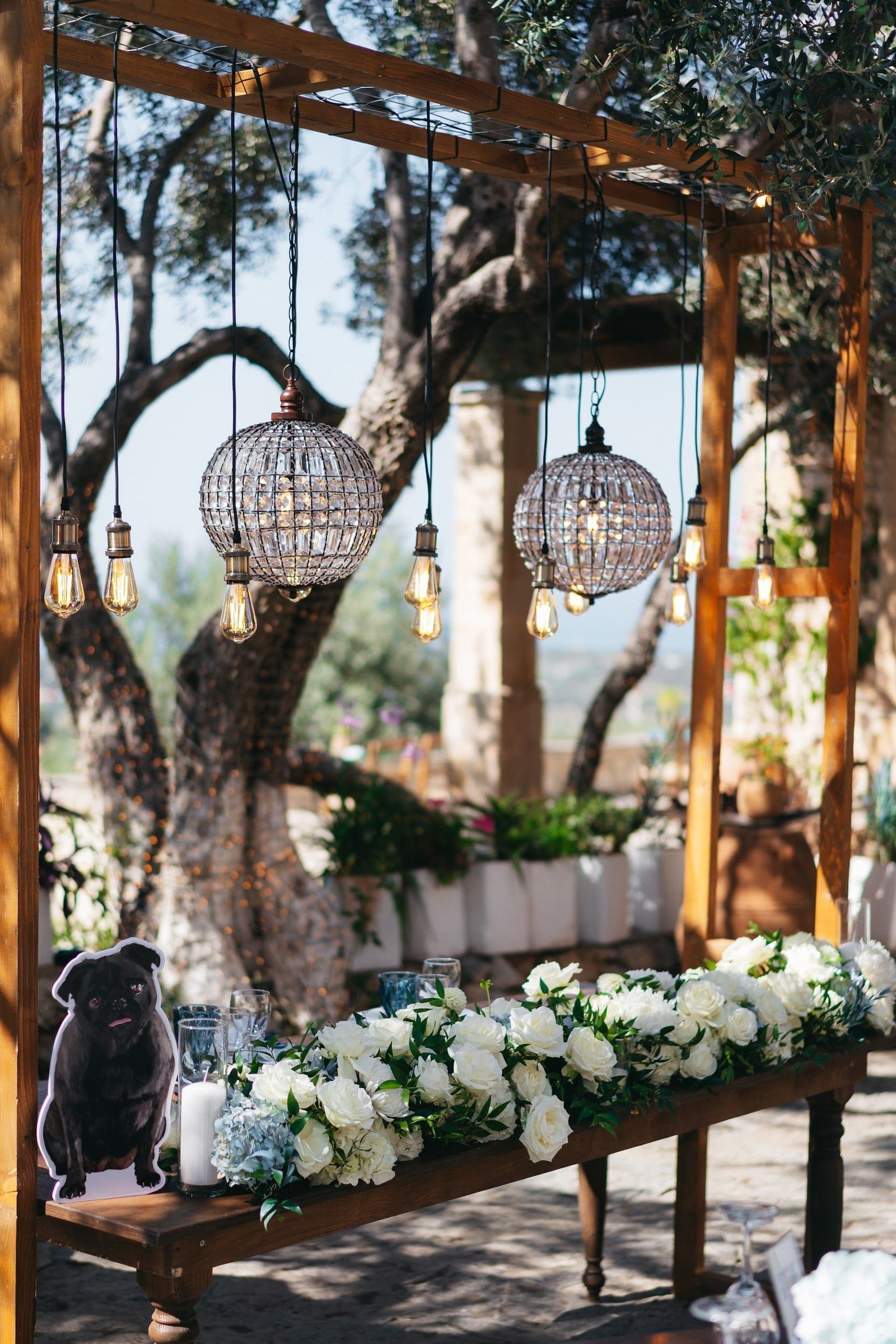 A long wooden table beneath a rustic timber pergola is dressed with a trailing runner of white garden roses, pale blue hydrangeas and green foliage, with white pillar candles and tall glasses set along it. Two large beaded crystal chandeliers and several bare Edison bulbs hang from the frame, and an old olive tree and stone pillar rise behind in soft afternoon light. This wedding table flower display at AgrecoFarms in Adele, Crete, Greece, has a warm, romantic, countryside feel.