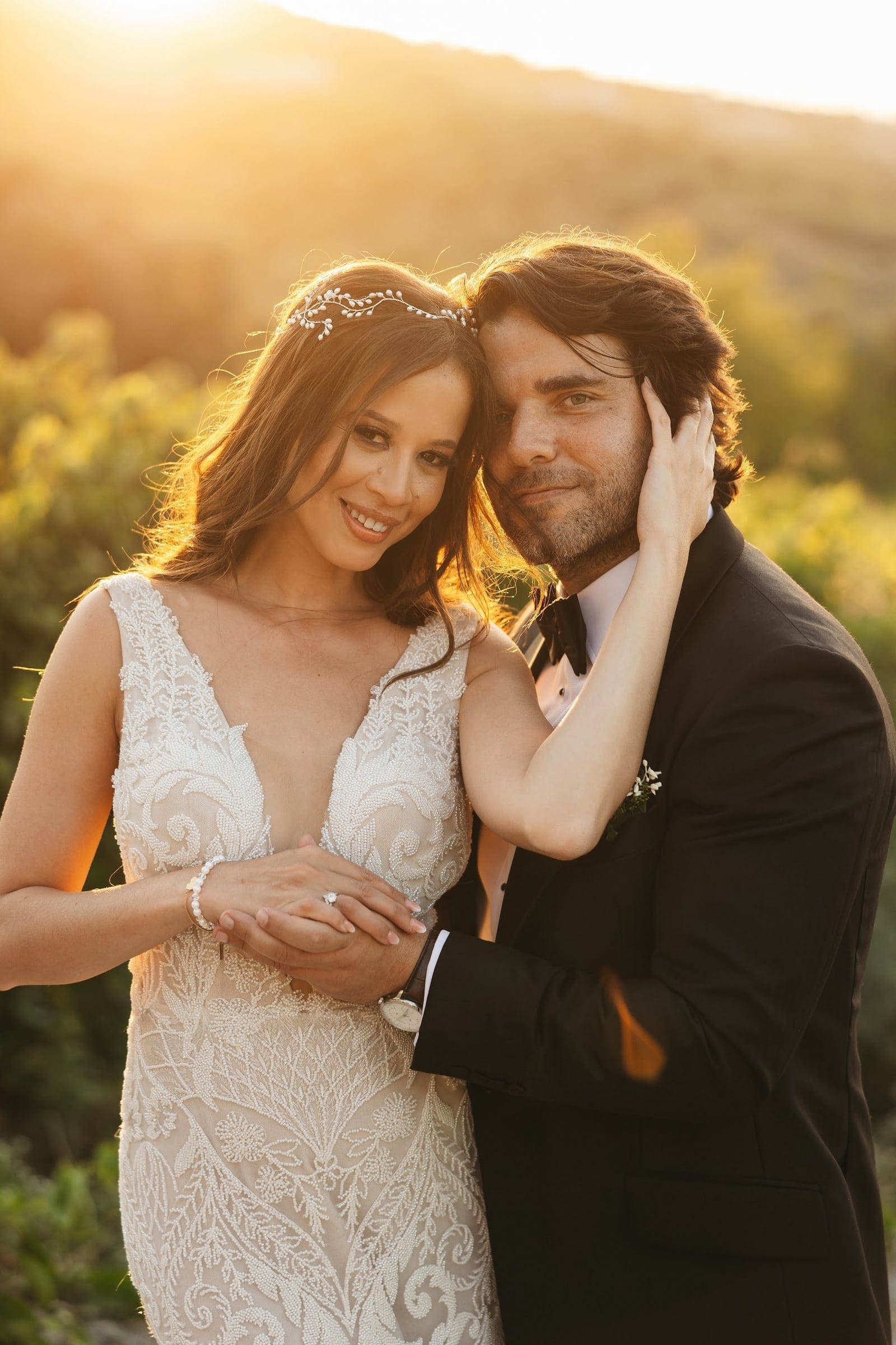 A bride and groom embrace in warm golden sunlight at Agreco Farm in Greece, with soft green foliage blurred behind them. The bride wears a beaded ivory gown with a plunging neckline and a pearl vine in her hair, while the groom wears a black tuxedo with a small white boutonniere, his head leaning against hers. Their hands are clasped at her waist, and the glowing backlight gives the moment a tender, romantic feel.
