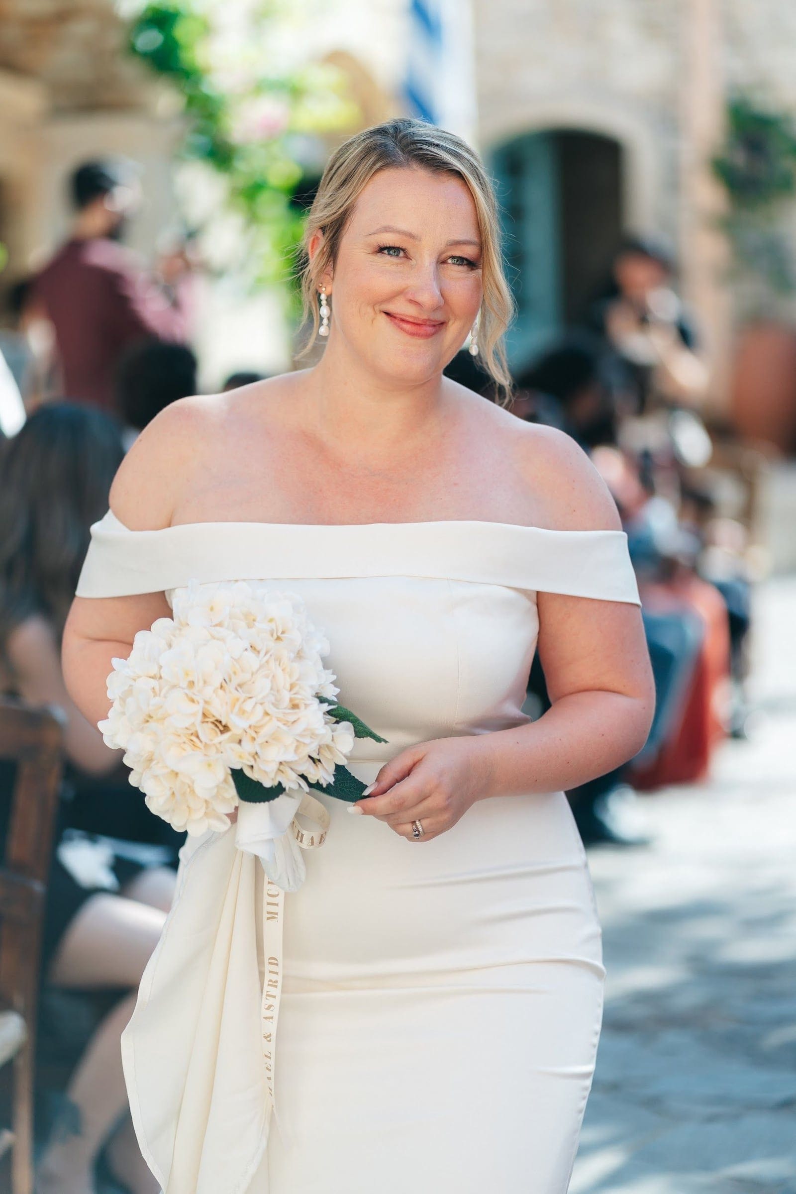 A bride in an ivory off-the-shoulder satin gown smiles softly while holding a bridal bouquet of cream hydrangeas with dark green leaves, its long ivory ribbon trailing down past her hip. Behind her, guests seated in rows and a stone-walled courtyard with a glimpse of green foliage blur into soft light, at a wedding at AgrecoFarms in Adele, Crete, Greece.