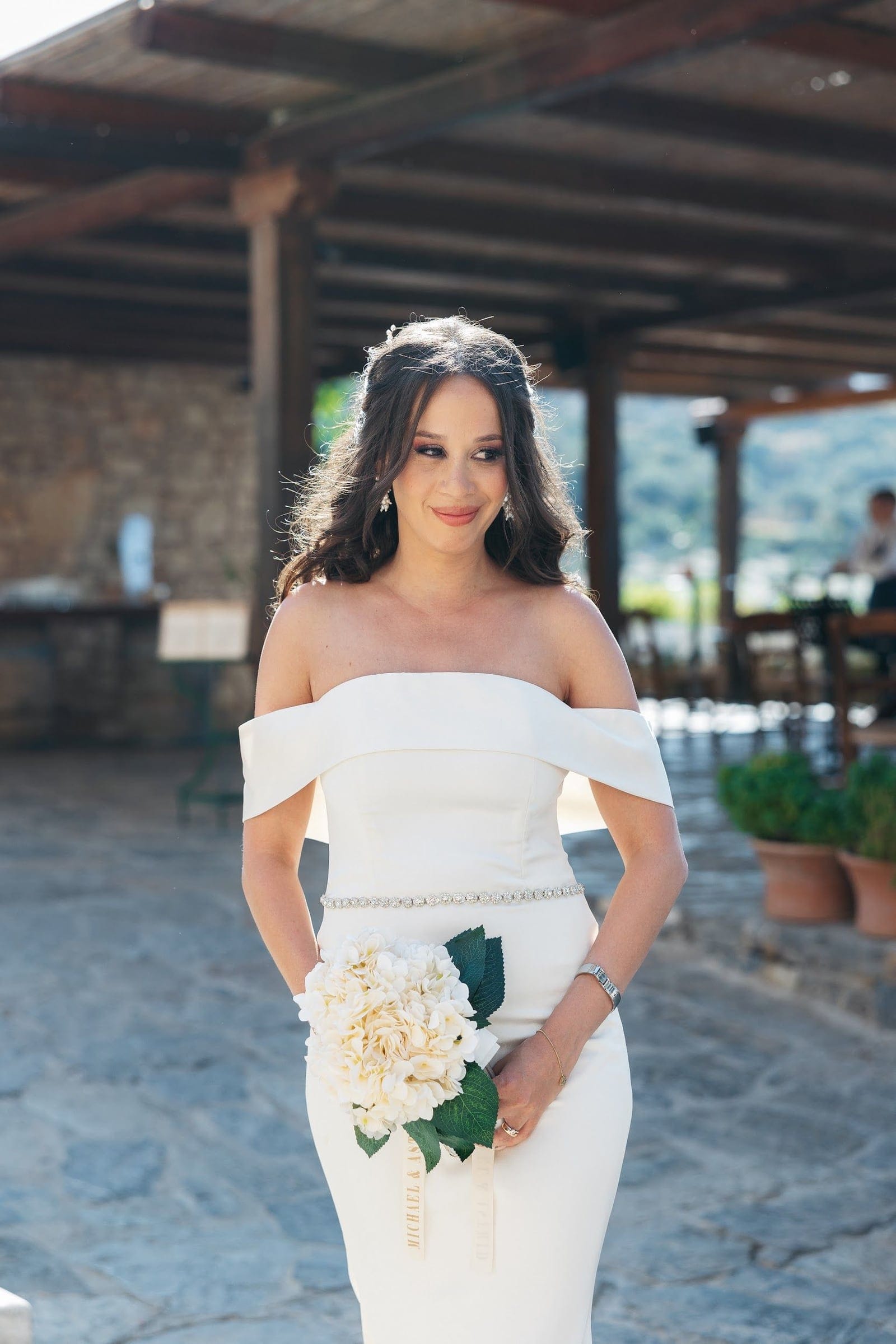 A bride in a fitted white off-shoulder gown with a jeweled waist belt stands on a stone terrace, holding a small bouquet of cream hydrangeas with dark green leaves, its gold ribbon trailing. Her dark hair is softly waved and pinned back, and she glances to one side with a gentle smile. Behind her, a timber-beamed pergola and stone wall frame a view of green hills, a wedding bouquet photographed at AgrecoFarms in Adele, Crete, Greece.