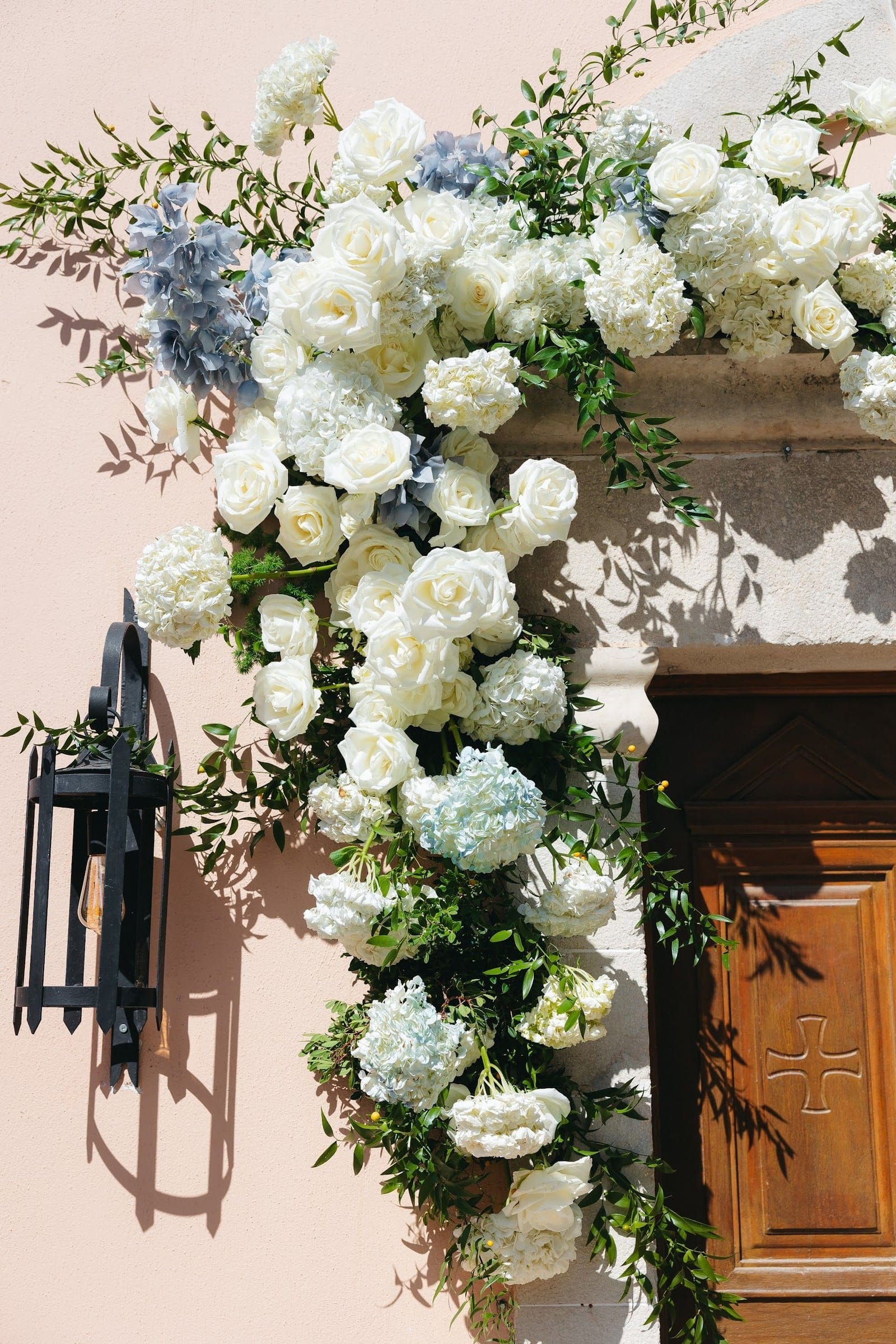 A cascading floral installation of white garden roses, white hydrangeas and dusty blue hydrangeas, trailing with green foliage down a pale pink wall beside a carved wooden door, at AgrecoFarms in Adele, Crete, Greece. The flowers form a lush arc over the stone doorway and spill downward toward a black iron lantern on the left, set in warm sunlight that casts soft shadows across the blooms. This wedding flower display for Andria and Alan feels fresh, romantic and elegant.