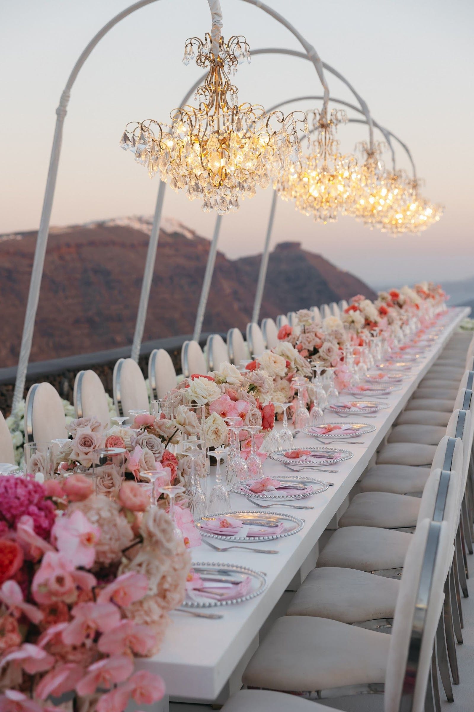 A long white banquet table at dusk on a clifftop terrace at Aenaon Villas in Santorini, Greece, set for a wedding dinner. Lush arrangements of blush roses, peach garden roses, pink orchids and cream hydrangea-like blooms run down its length, with pink-edged plates and crystal glasses in between. Three arched frames hang with glittering crystal chandeliers above, and beyond them the rust-coloured caldera cliffs and pale pink sky fade into the sea.