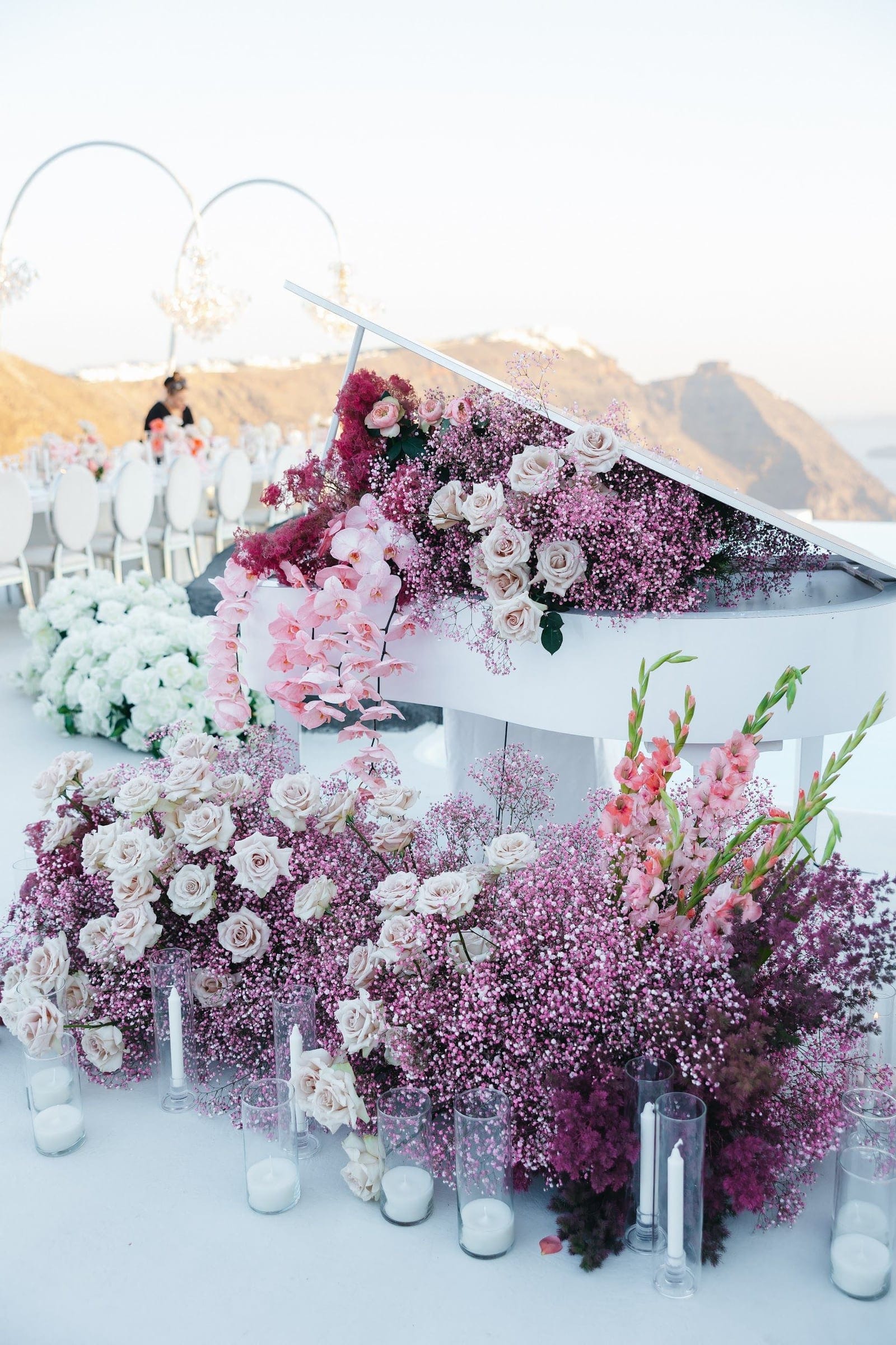 A white grand piano draped in cascades of pink gypsophila, blush garden roses and trailing pink orchids, with more blush roses and tall pink gladioli spilling across the floor around it. Glass hurricane candles line the base, and beyond, white chairs and an arch overlook the caldera at Aenaon Villas in Santorini, Greece, where this wedding flower installation sits on a sunlit terrace.