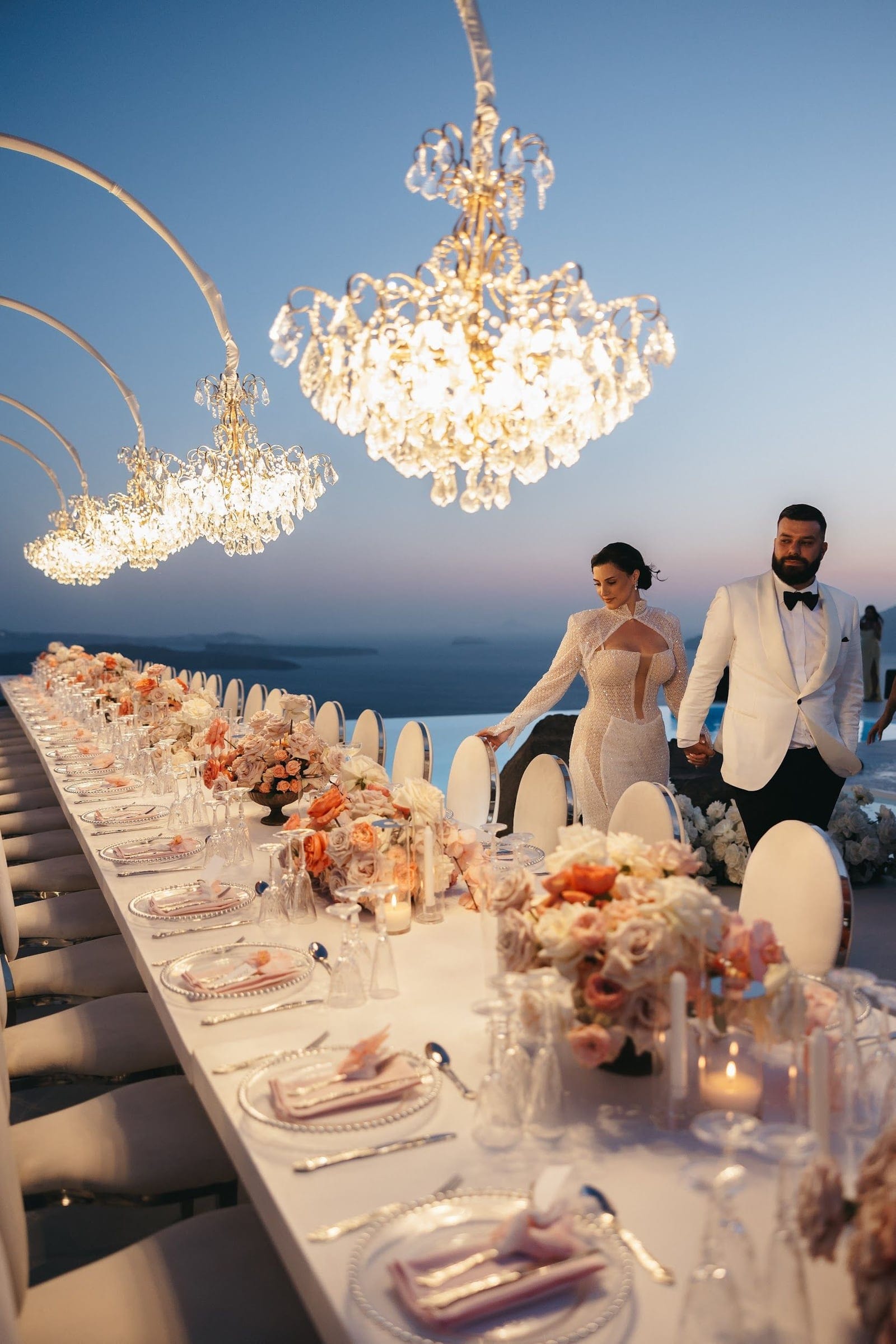A long white dining table stretches toward the sea, dressed with blush, peach and cream roses, orange ranunculus and candles in glass, with crystal chandeliers glowing above it at dusk. Beyond, a bride in a sheer, pearl-studded gown and a groom in a white dinner jacket and black bow tie walk hand in hand along the edge of the terrace, with the deep blue Aegean and dusky sky behind them. This is a wedding reception table with flowers at Aenaon Villas in Santorini, Greece.